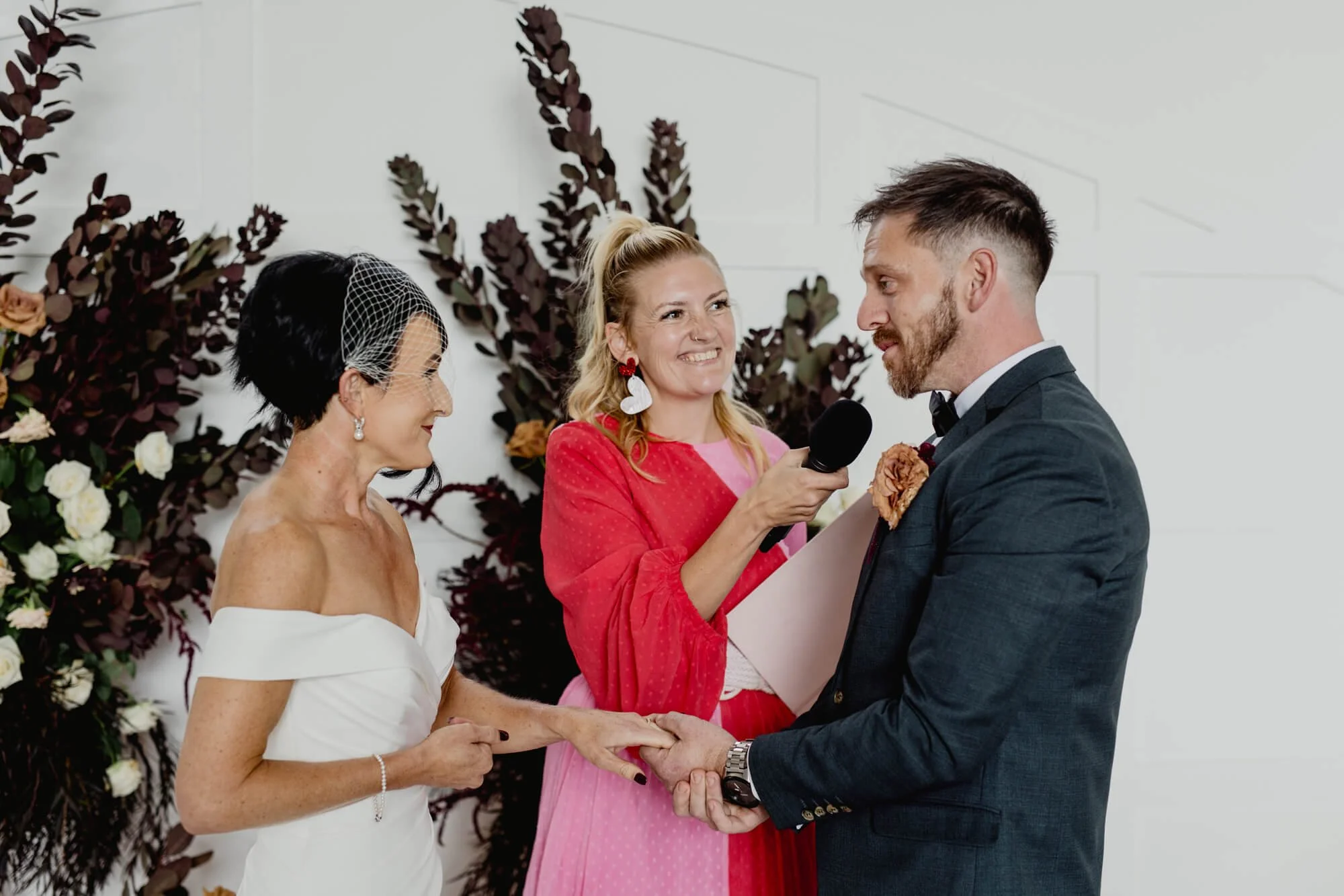 Bride and groom holding hands during wedding ceremony, officiant smiling, with floral and foliage backdrop.