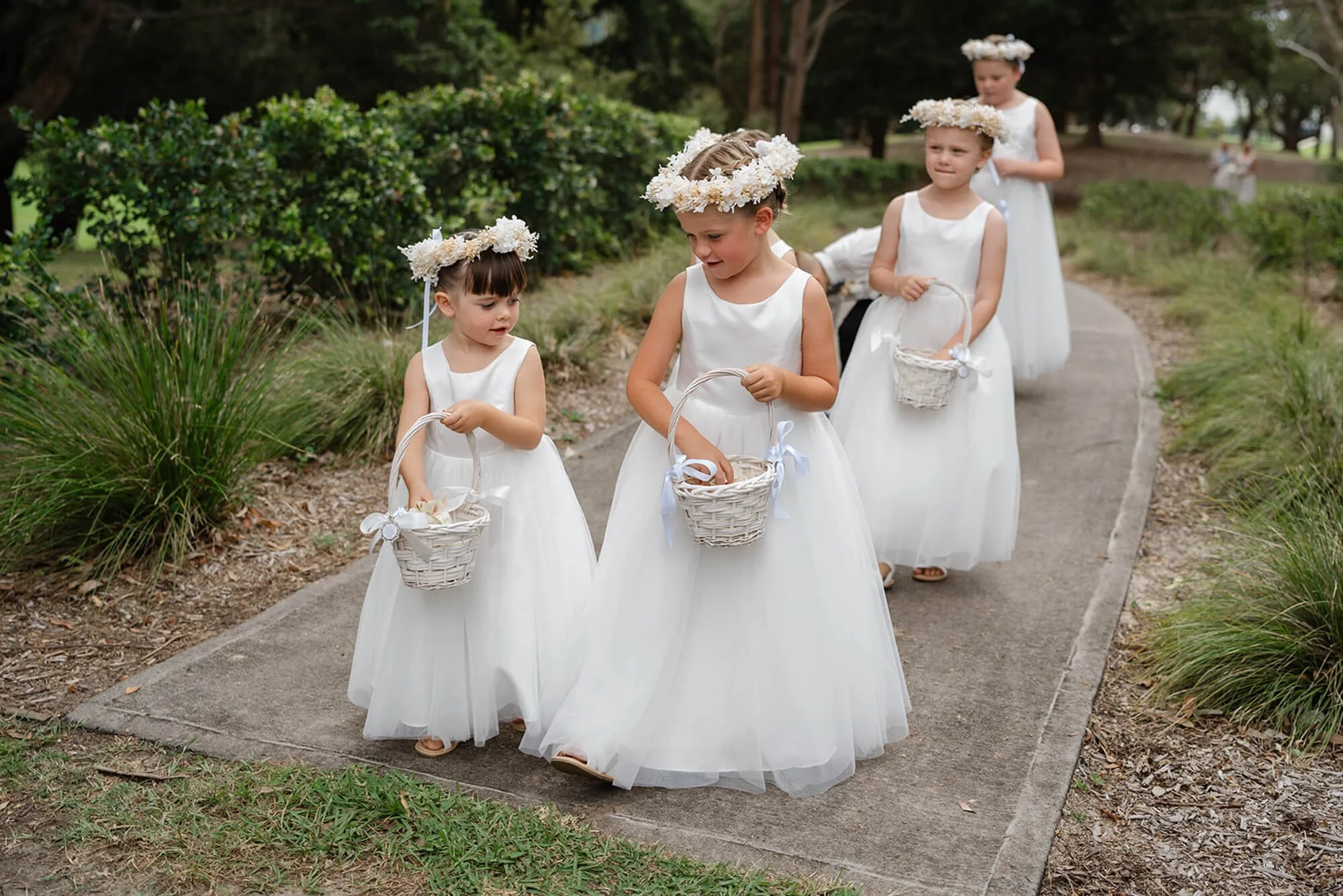Young girls dressed in white dresses with flower crowns, carrying baskets, walking along a garden path during a wedding or special event.