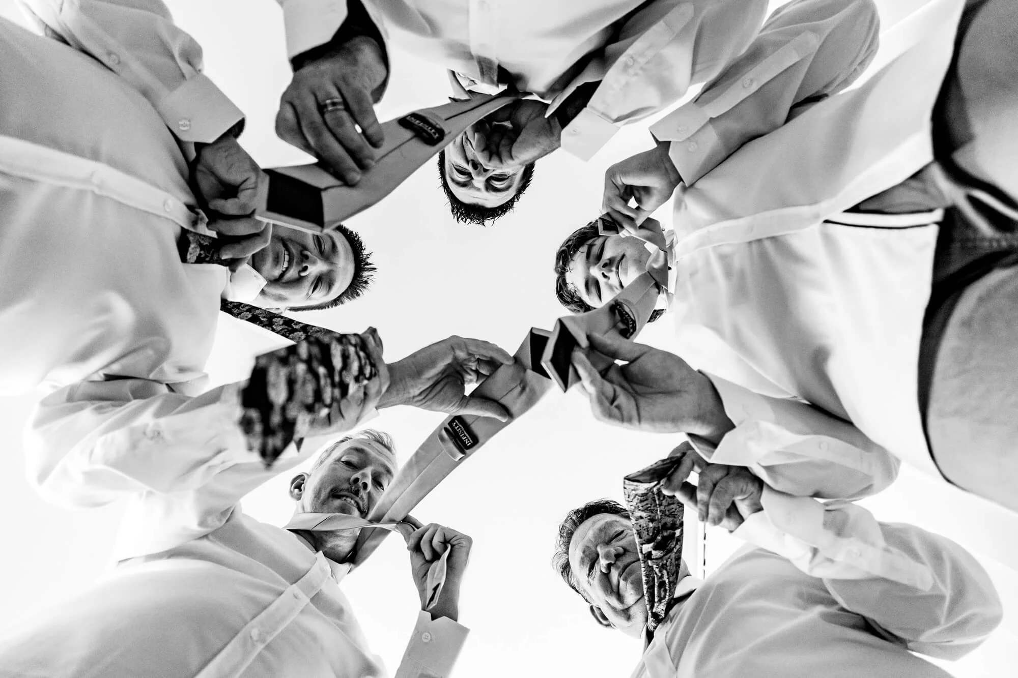 Six men in white shirts are standing in a circle, looking down at the camera, holding sports trophies, with some smiling and others serious.