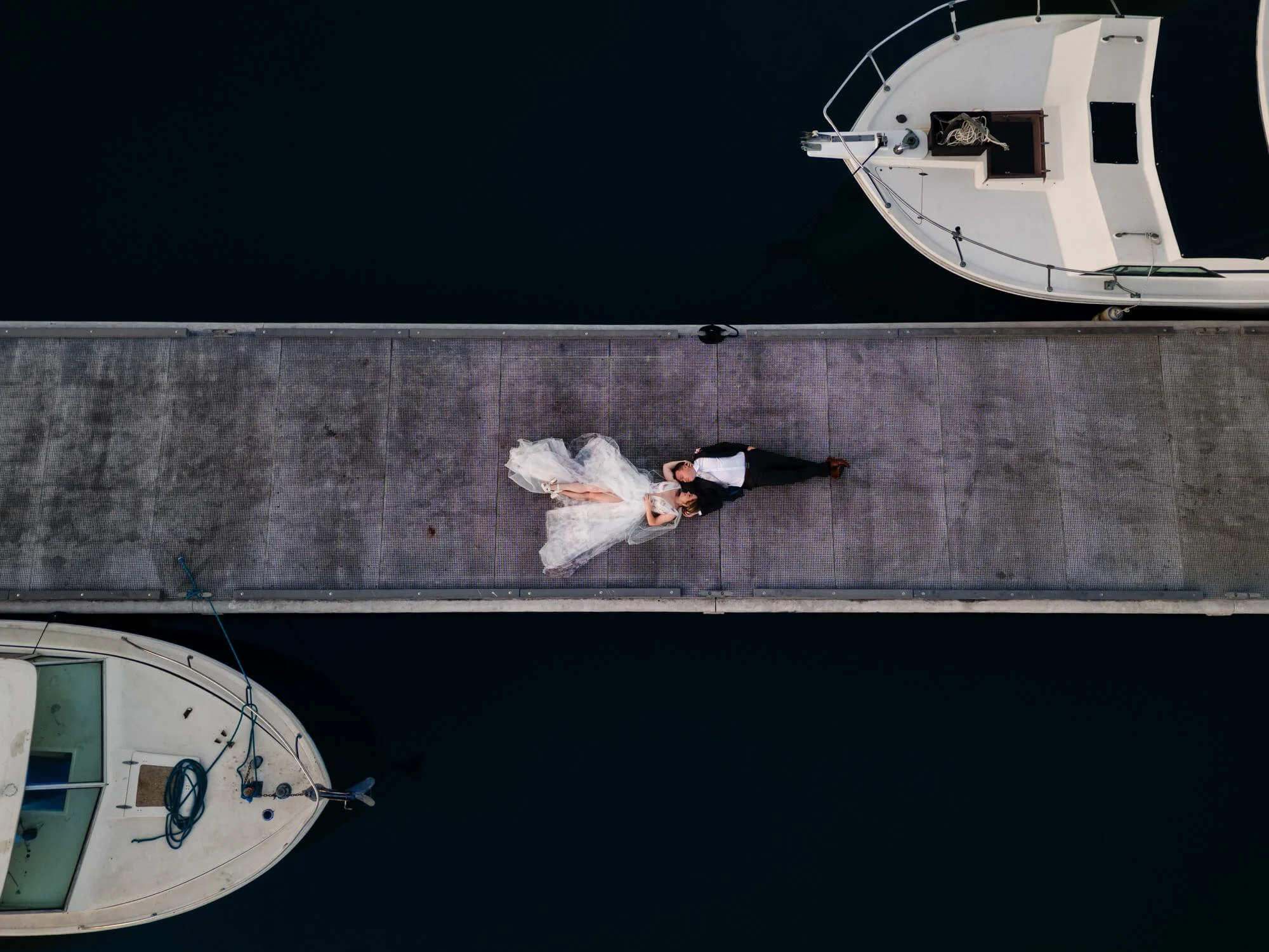 A bride and groom are lying on a dock by the water, looking up at the camera, with a boat on each side of the dock.