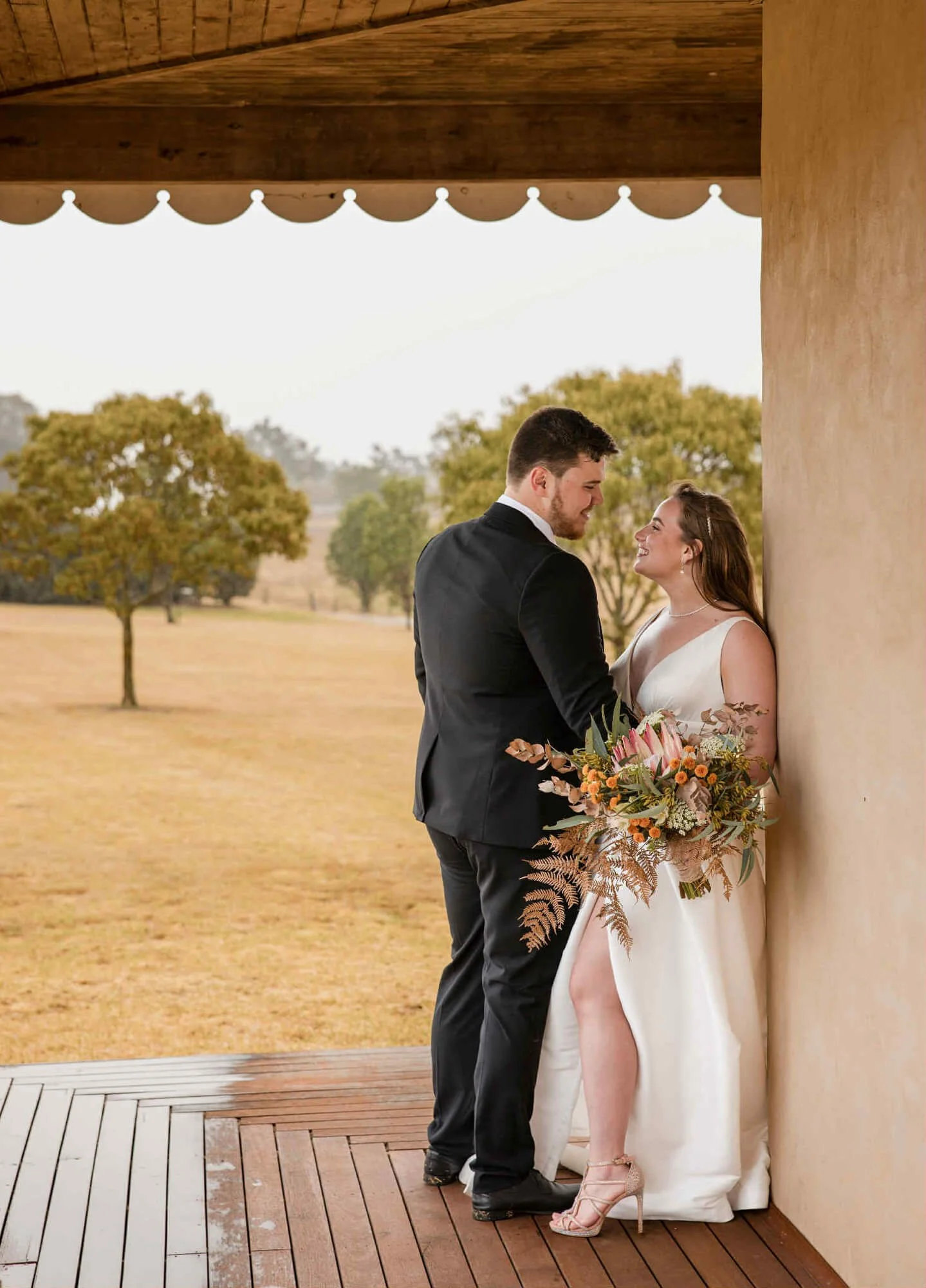 A couple in wedding attire standing closely together under a porch, with a scenic outdoor background of trees and open field.