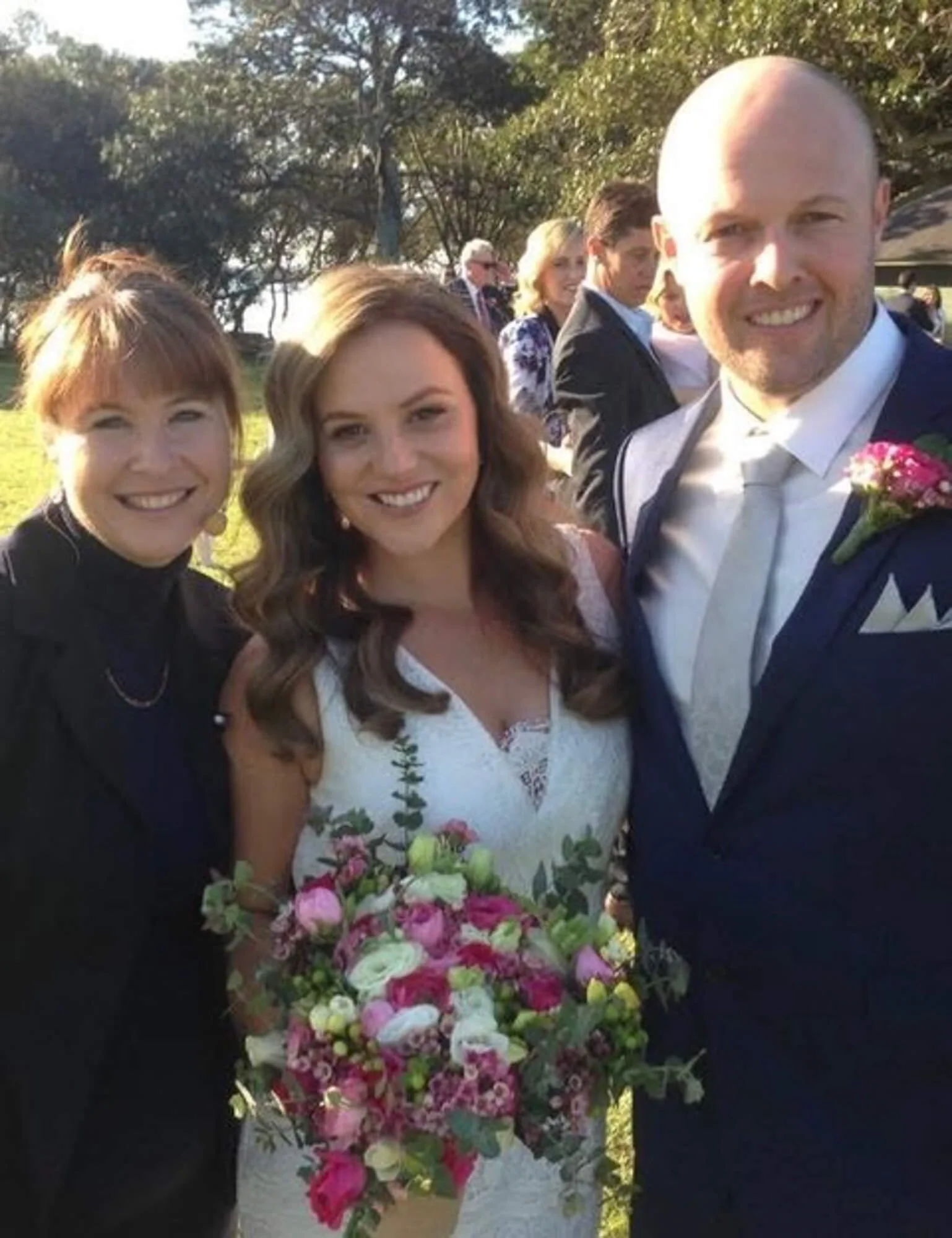 A group of four people dressed in formal attire at an outdoor event, with greenery and other guests in the background, celebrating a wedding.
