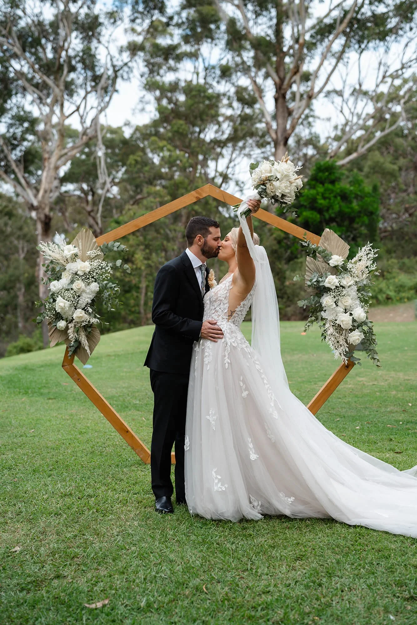 A bride and groom kiss at their outdoor wedding ceremony, standing inside a geometric wooden arch decorated with white flowers and greenery, on a grassy field with trees in the background.