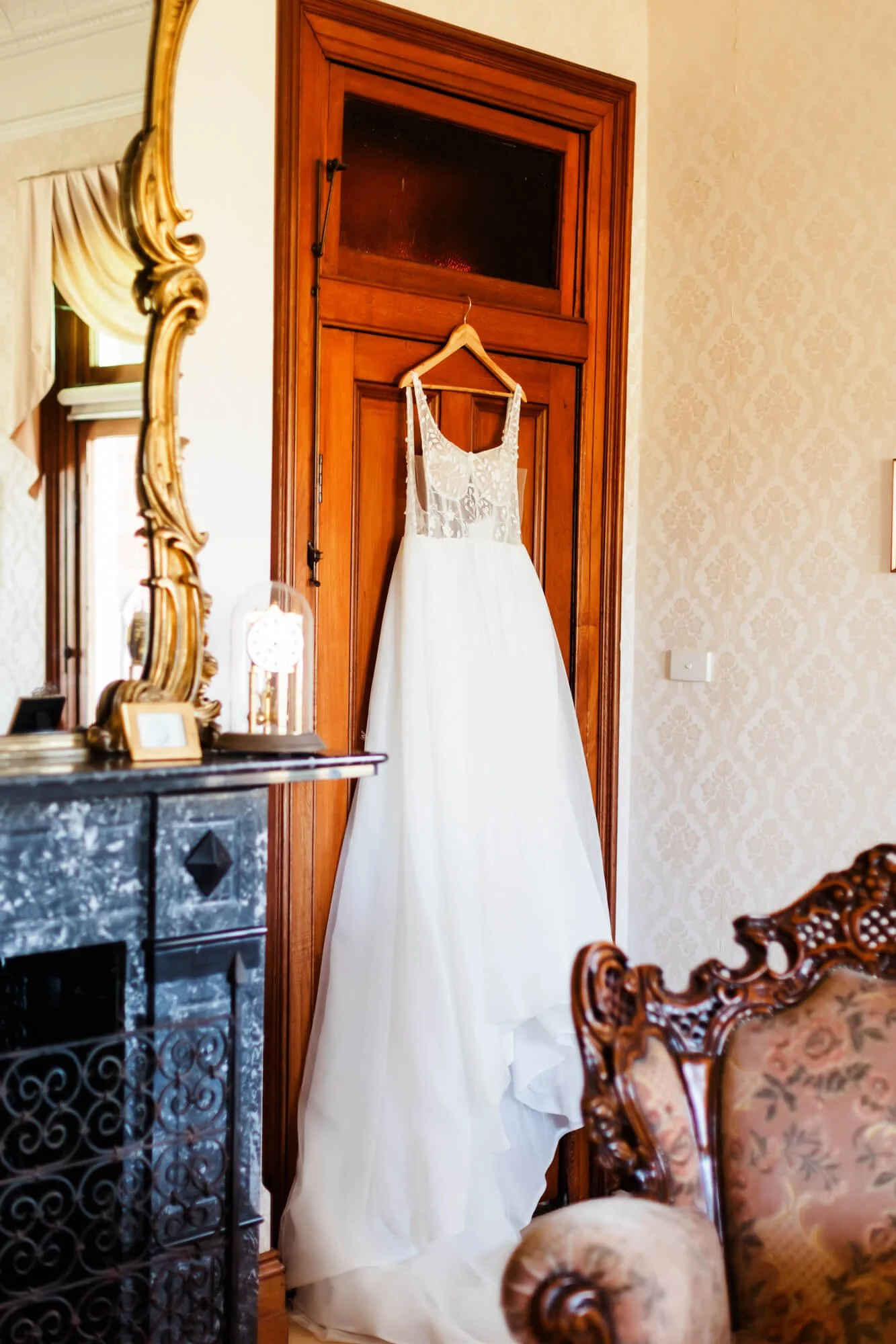 A white wedding dress hangs on a wooden door in a decorated room, with ornate furniture and a large mirror visible.
