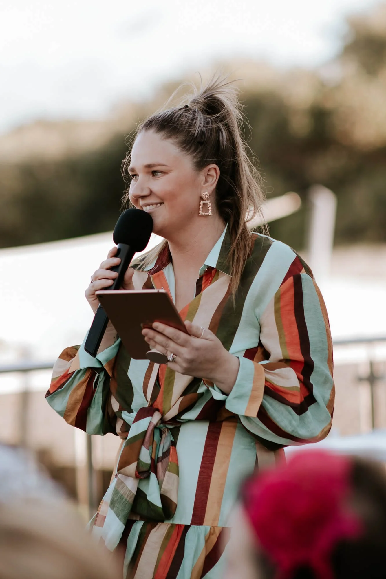 Woman with long brown hair in a ponytail, holding a microphone and a tablet, smiling, wearing a colorful striped blouse and statement earrings, outdoors during daytime.