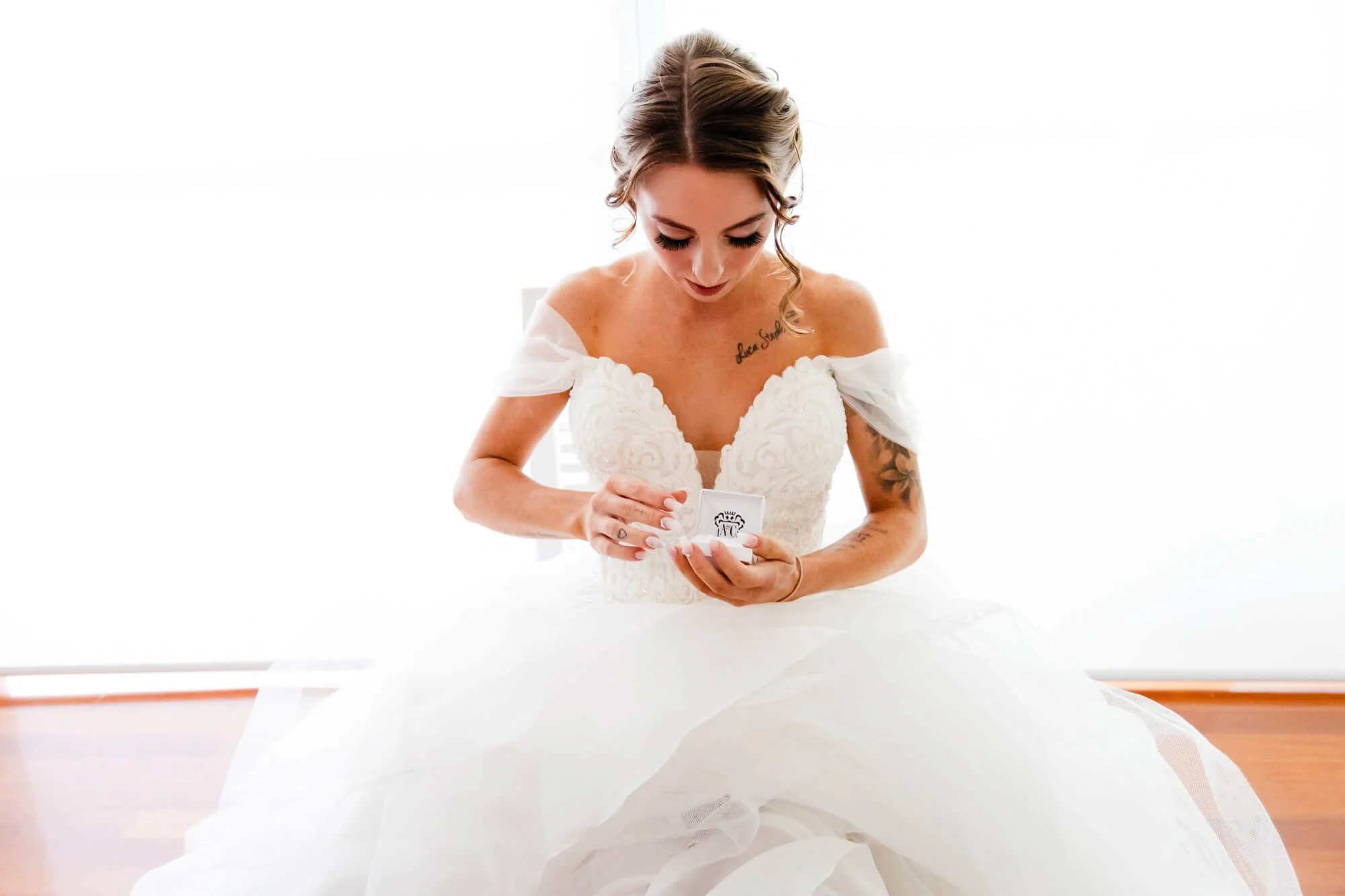 A bride in a white wedding dress with off-the-shoulder sleeves sits on the floor, looking down at a small box she is opening, in a brightly lit room with white walls.