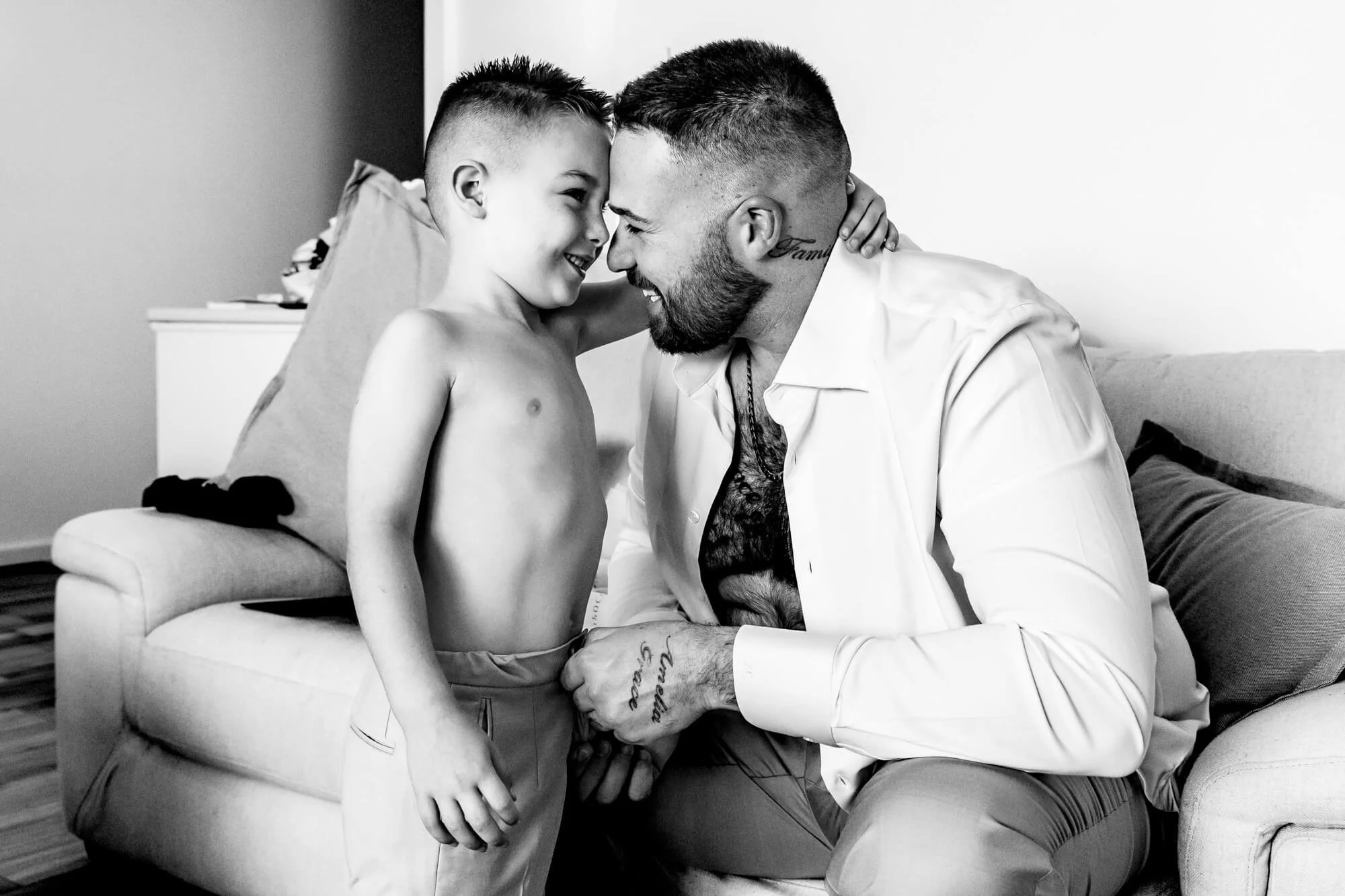 A shirtless young boy and a man with a beard and tattoos are touching foreheads and smiling at each other in a living room.