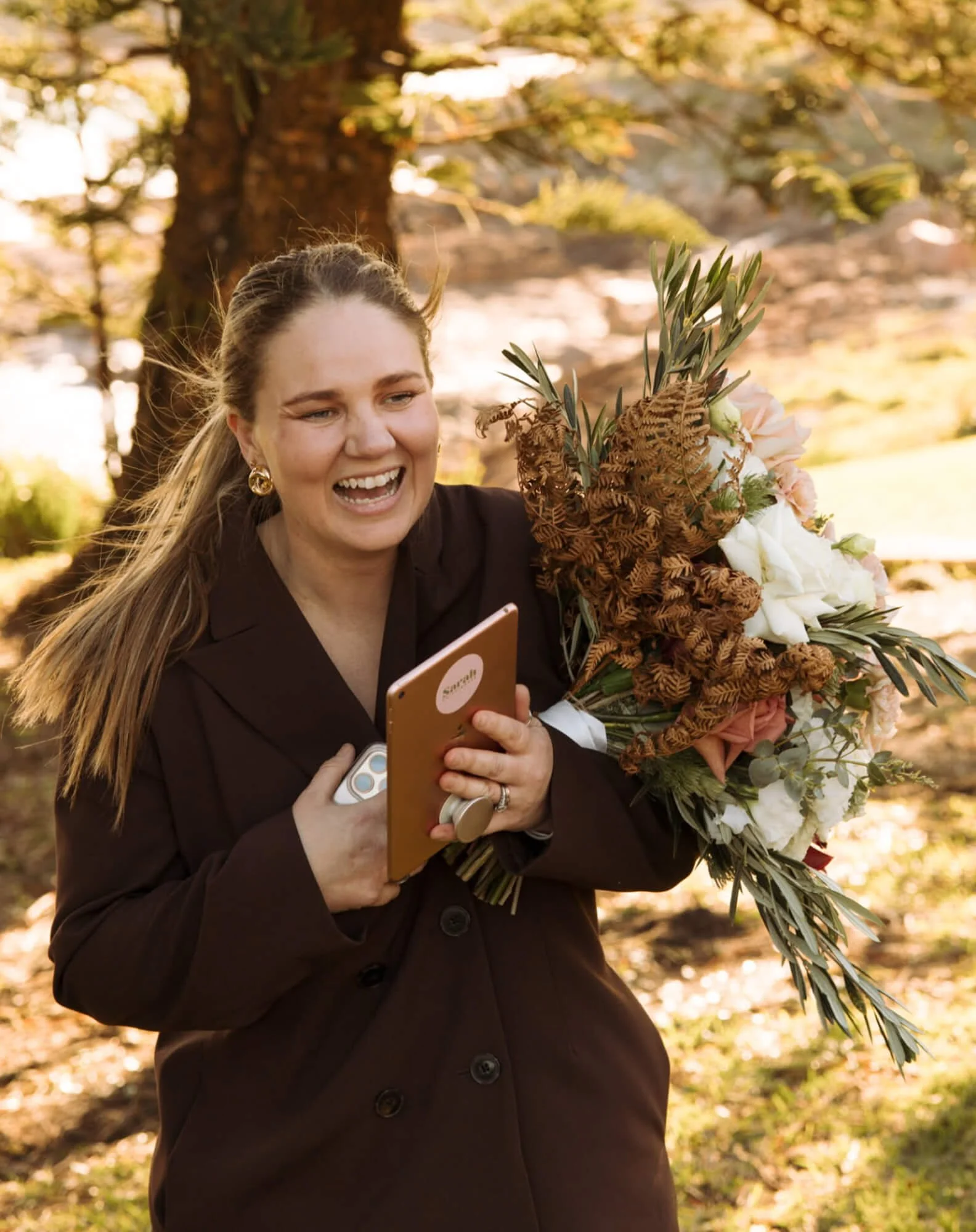A woman holding a large bouquet of flowers and looking at her phone outdoors near a tree, smiling and appearing to be happy.