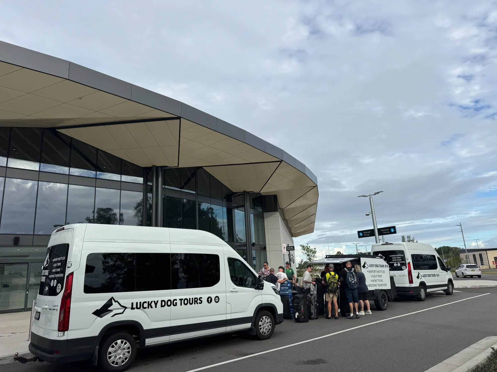 People gathering at an airport with two white shuttle vans labeled "Lucky Dog Tours," parked in front of a modern building with a curved roof and glass windows.