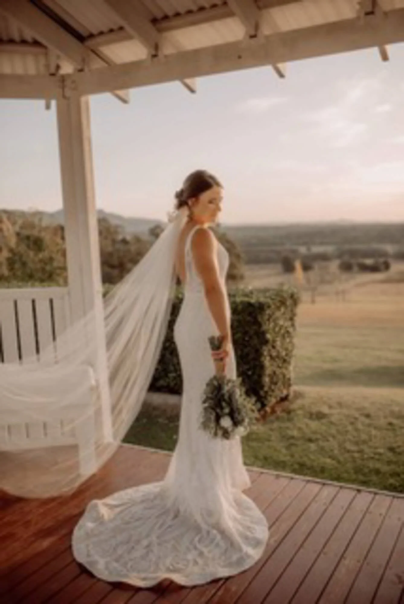 A bride in a white wedding dress holding a bouquet stands on a wooden deck, looking at a scenic landscape during sunset.