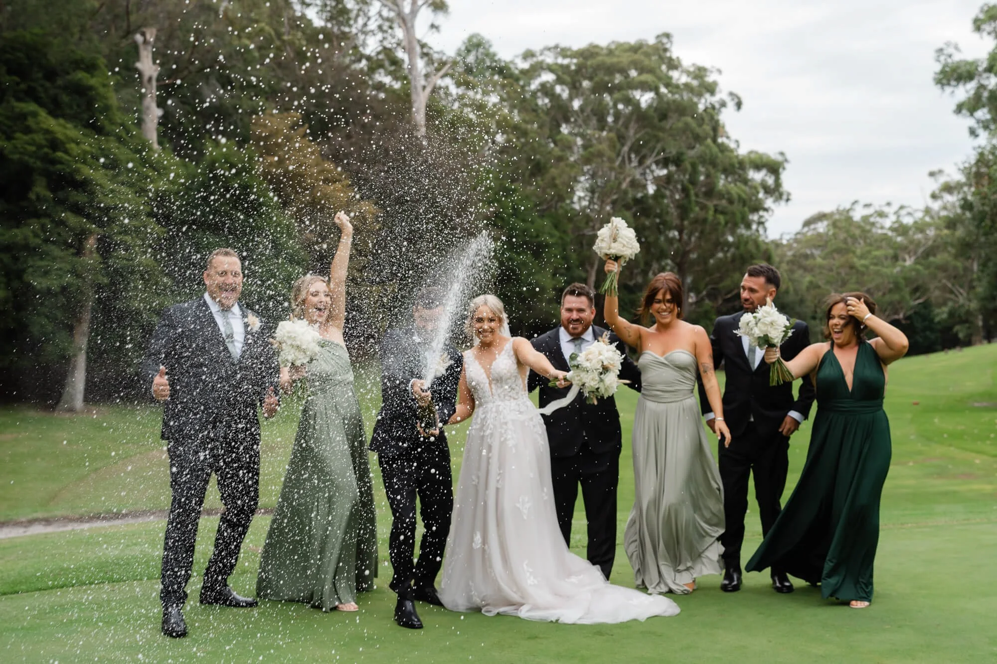 Group of people celebrating at a wedding outdoors, with a bride in a white gown, other guests in formal dresses and suits, some holding bouquets, while spraying champagne and smiling.