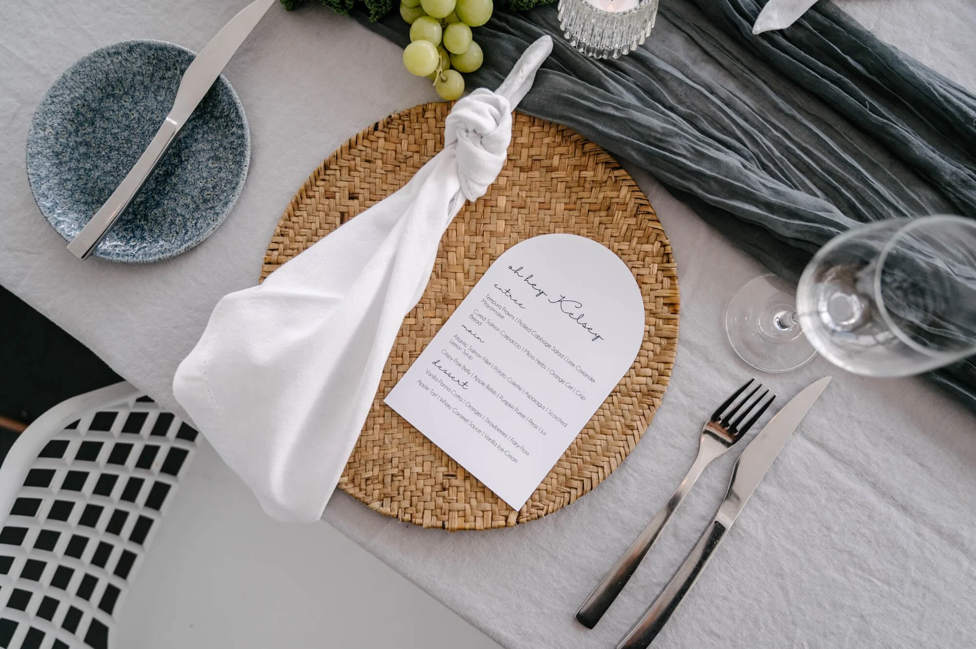 Elegant table setting with a woven placemat, a white napkin tied with a knot, a menu card, blue and clear glassware, a blue patterned plate with silverware, a bunch of green grapes, and a dark gray cloth runner.