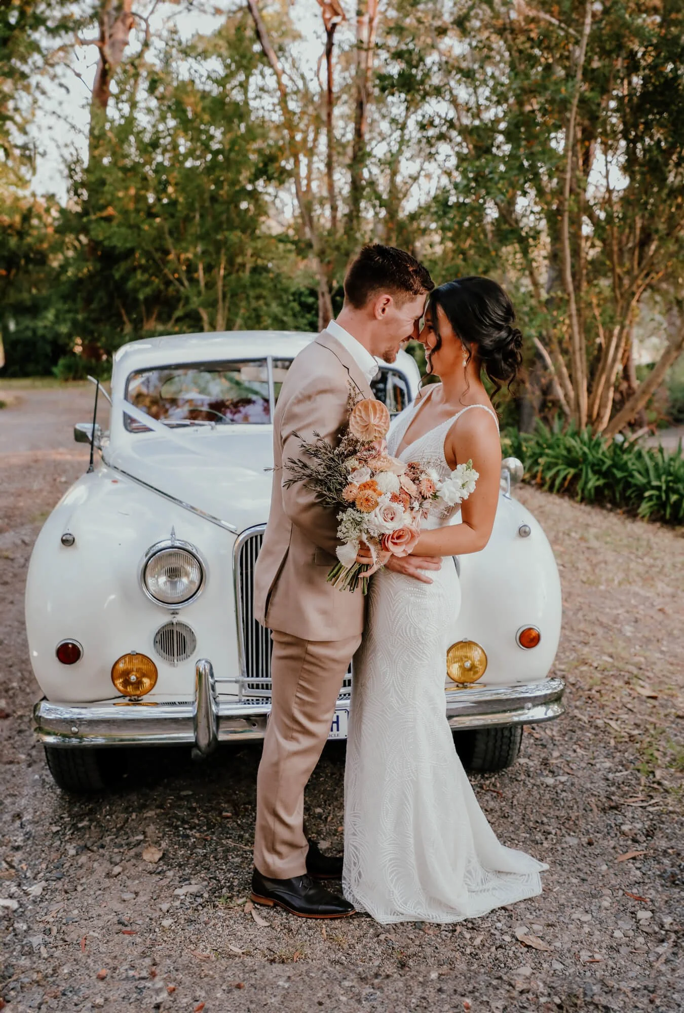A newlywed couple standing close together in front of a vintage white car, smiling and touching foreheads, outdoors surrounded by trees during sunset, with the bride holding a bouquet of pink, white, and orange flowers.