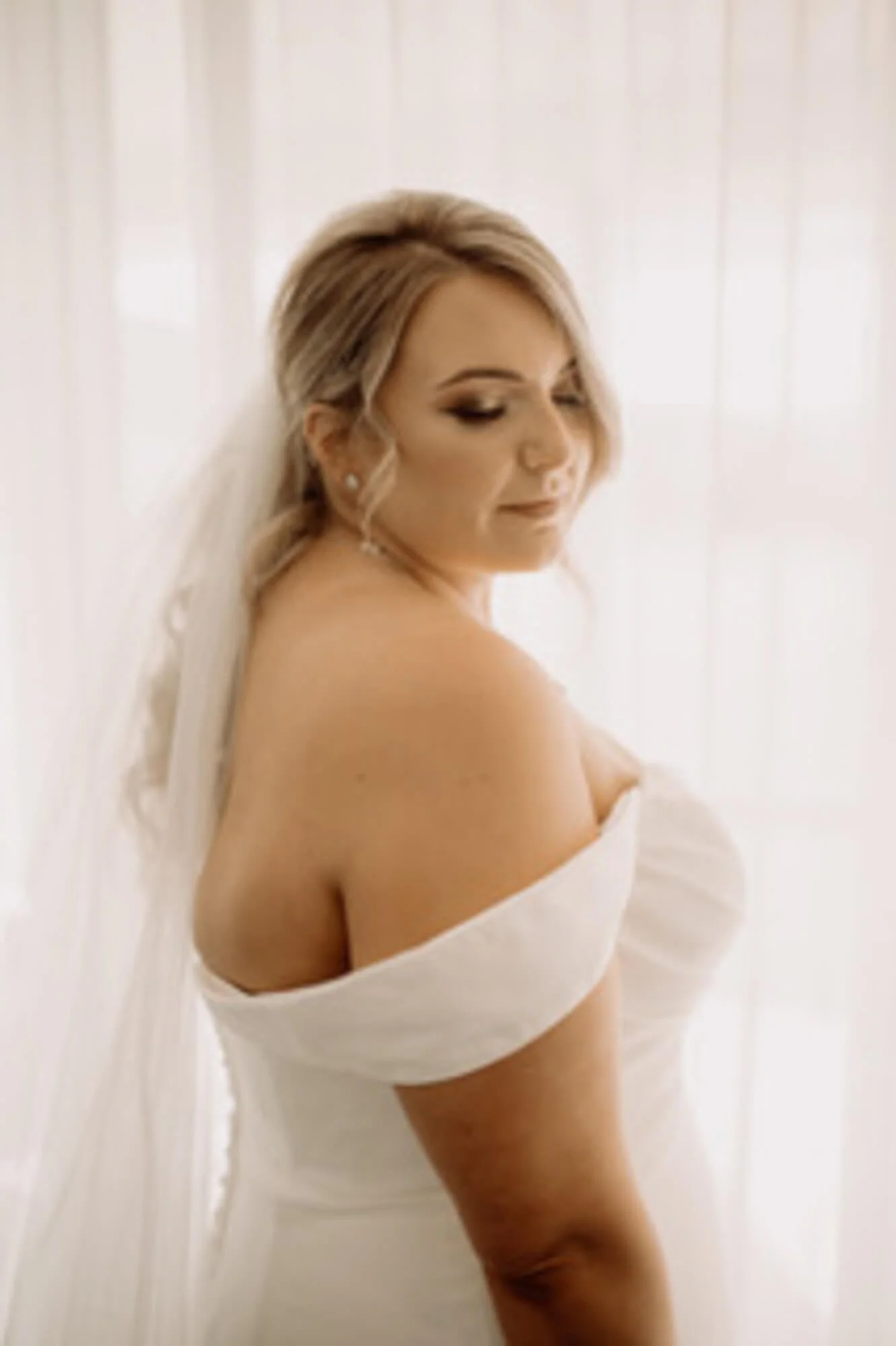 A woman with blond hair styled in soft waves, wearing makeup with dark eyeshadow, earrings, and a white off-the-shoulder dress, standing against a white background with soft lighting.