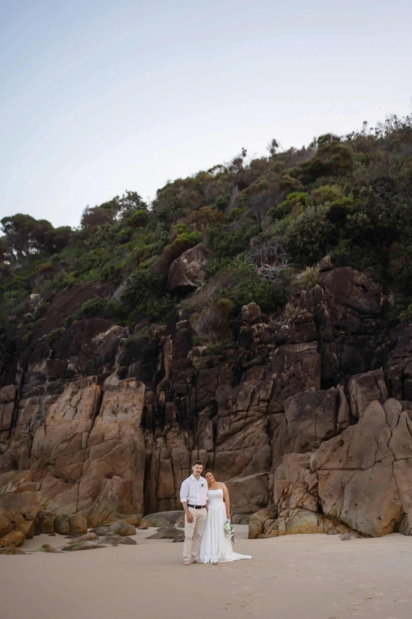 A bride and groom standing on a sandy beach with large rocks and cliffside with greenery behind them.