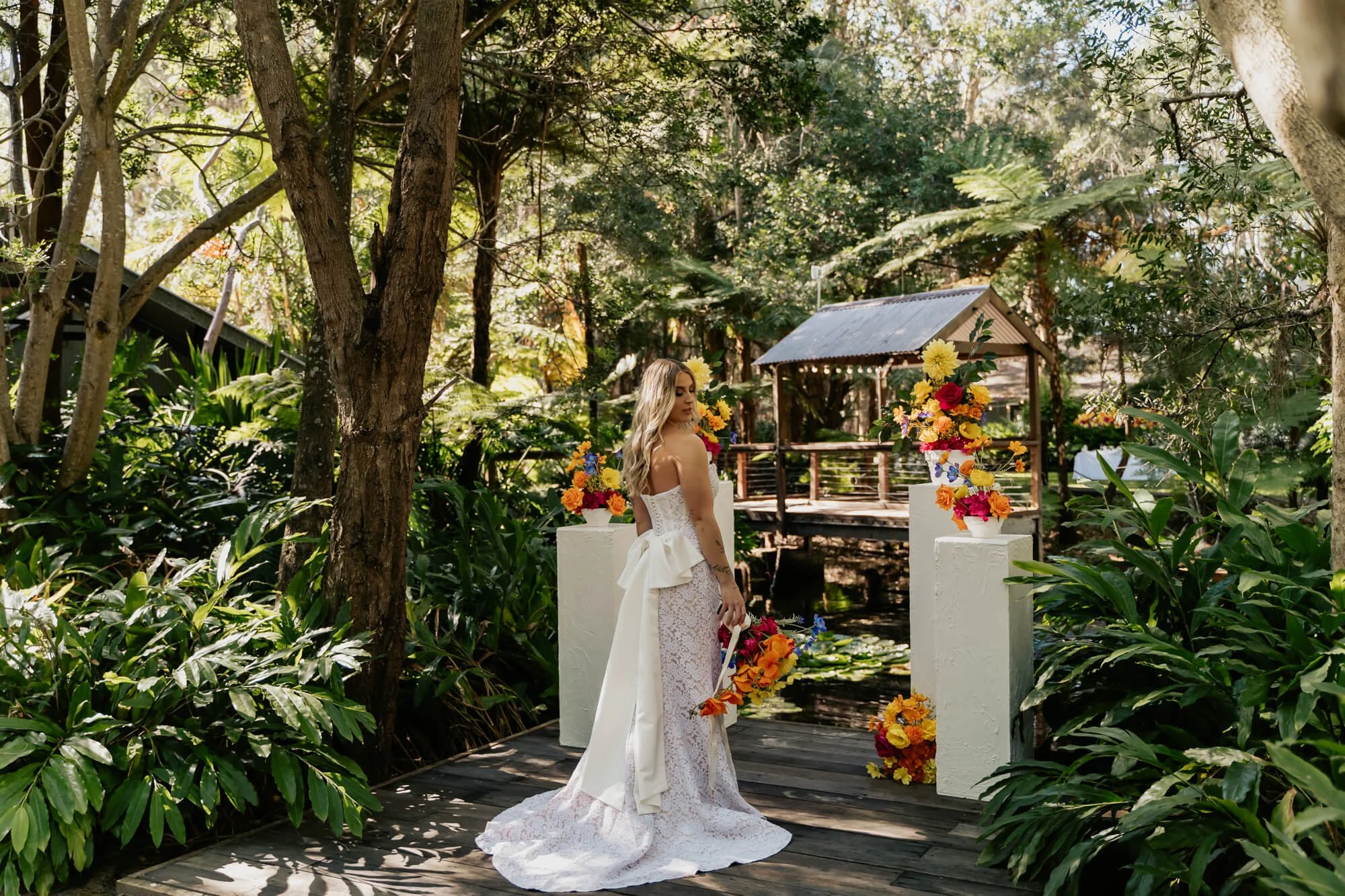 A woman in a wedding dress holding a bouquet stands on a wooden path in a lush, green garden with trees and plants. There are two white pedestals with colorful flower arrangements and a small wooden gazebo in the background, with sunlight filtering t