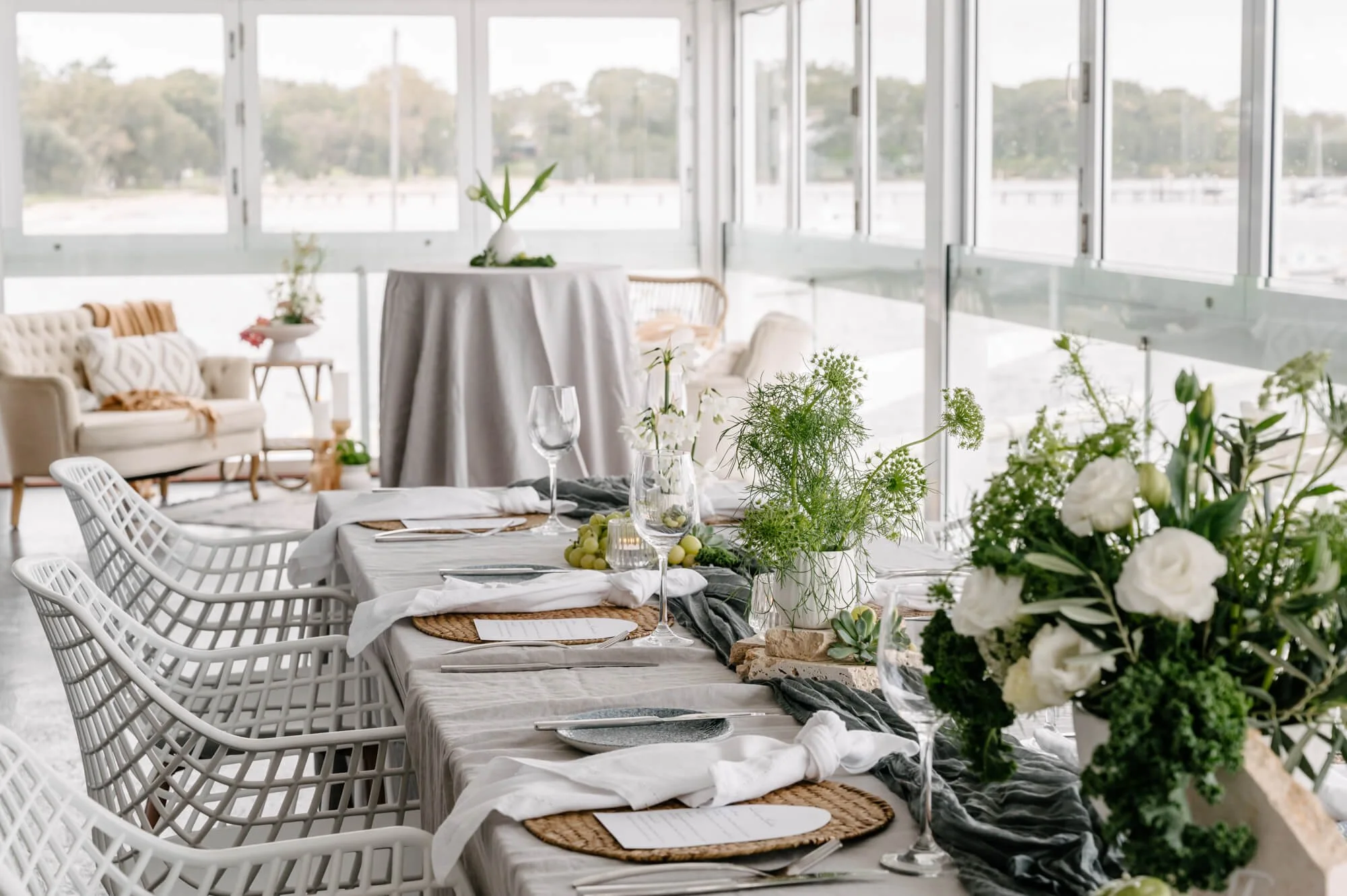 Elegant dining area with a long table set for a meal, decorated with white and green floral arrangements, wine glasses, and woven placemats. The room has large windows and a cozy seating area with a sofa in the background.
