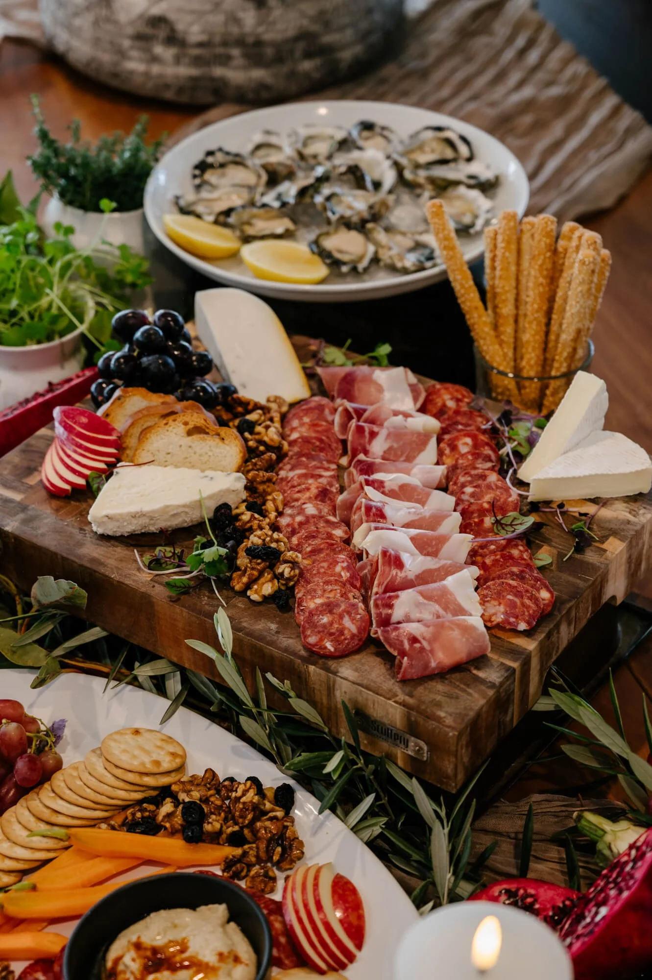 A charcuterie board with an assortment of cured meats, cheeses, crackers, and grapes, surrounded by fresh herbs and a spread of seafood and other snacks.