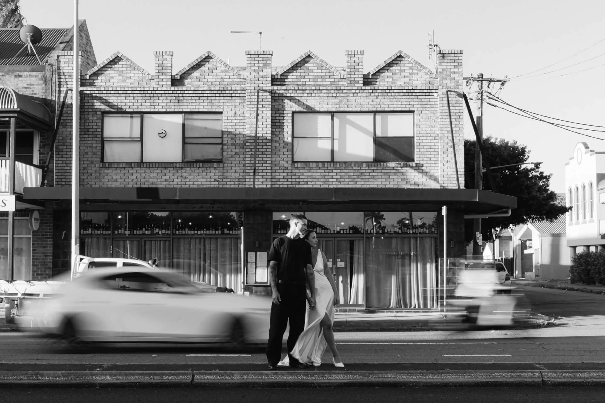 A black and white photo of a city street with a modern brick building in the background. Two people, a man and a woman, are walking hand-in-hand across the crosswalk in front of the building, with a moving car passing by.