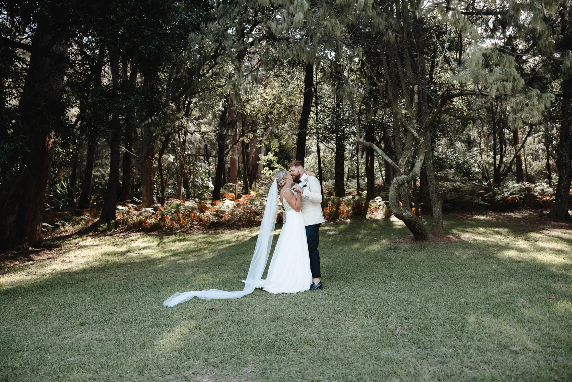 A bride and groom share a kiss in a forest clearing, surrounded by trees and foliage.