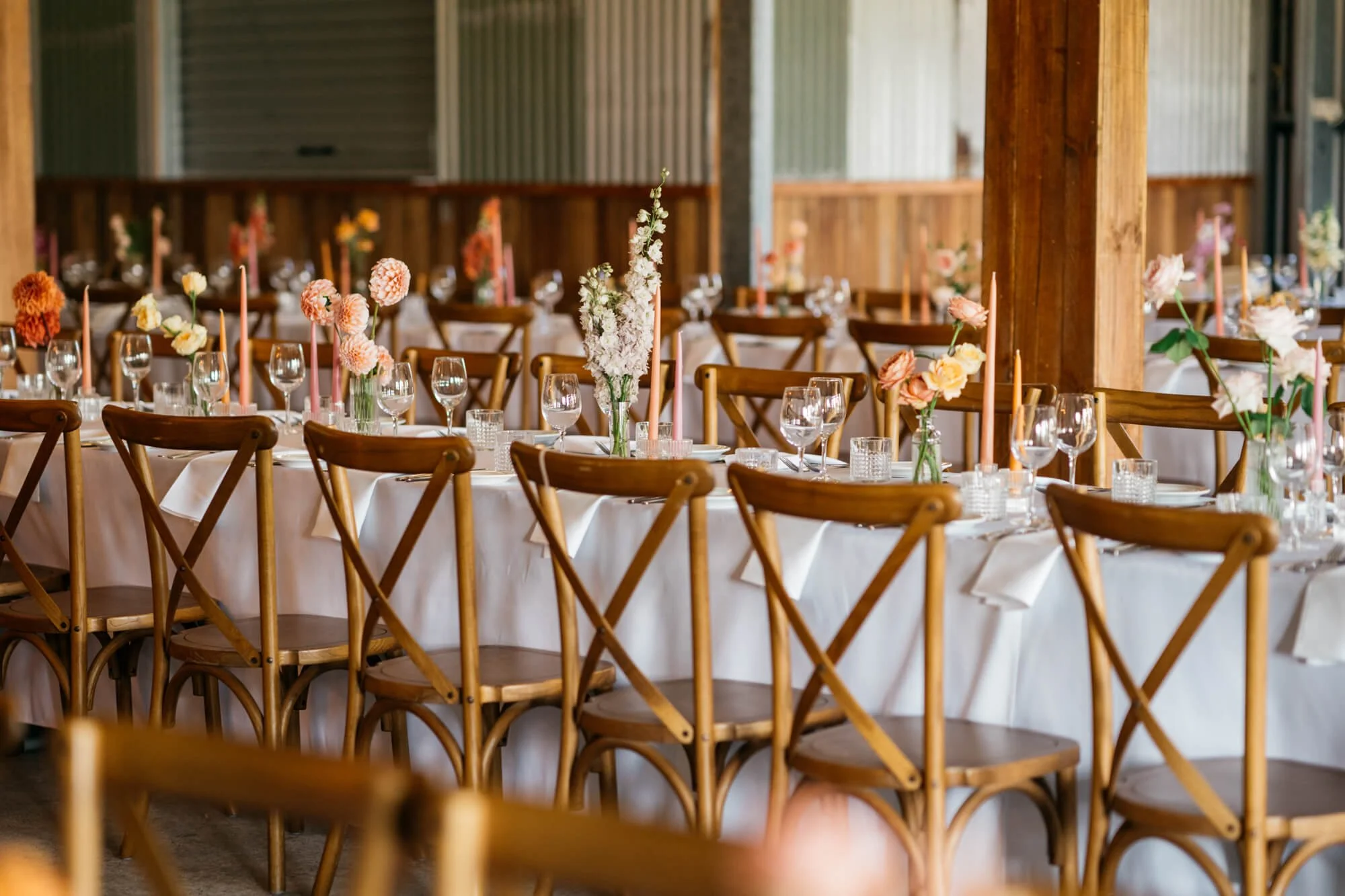 Decorated banquet table with floral arrangements, pink candles, wine glasses, and white tablecloths in a rustic setting.