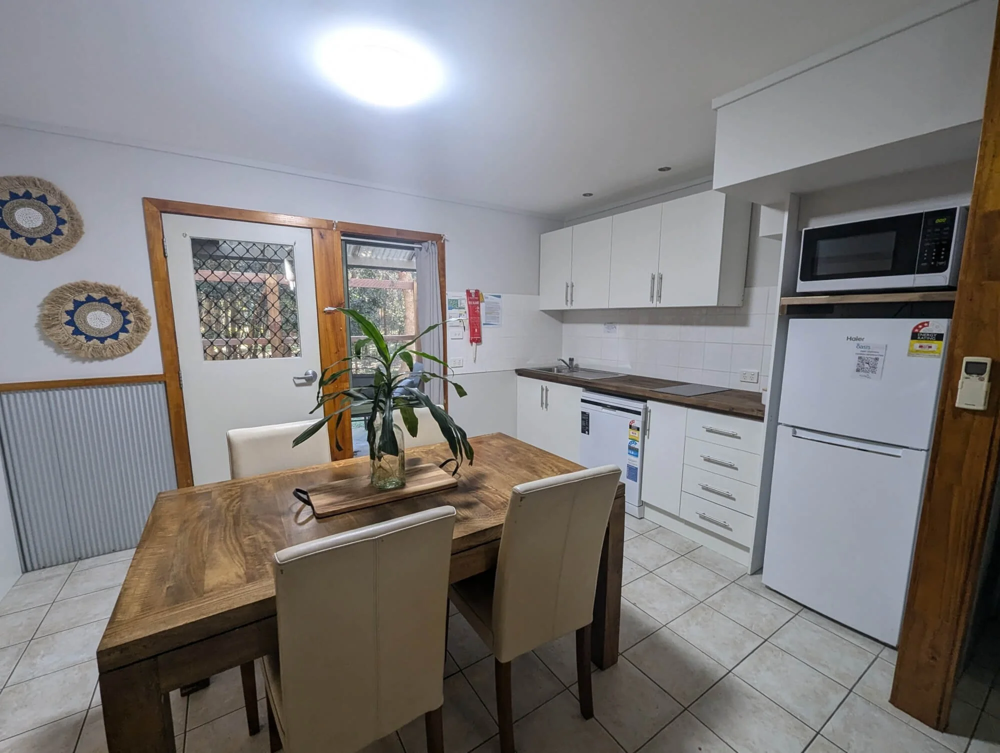 A kitchen with white cabinets, a wooden dining table with four beige chairs, a potted plant on the table, white tiled floor, and a door leading outside with a windowed door that has a security screen. Decor on the wall includes two wicker-style woven