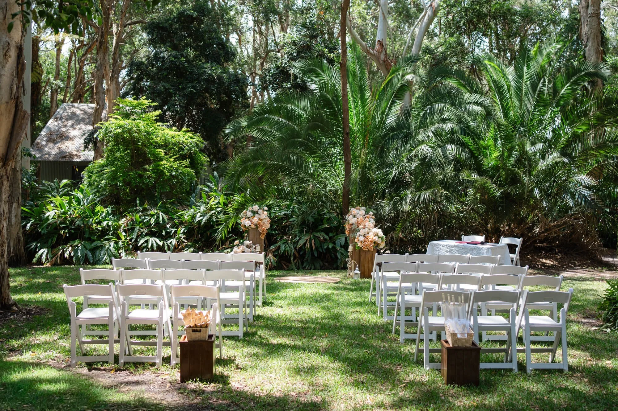 Outdoor wedding ceremony setup on grassy area with white chairs arranged in rows, floral decorations, and a table at the side, surrounded by lush green trees and plants.