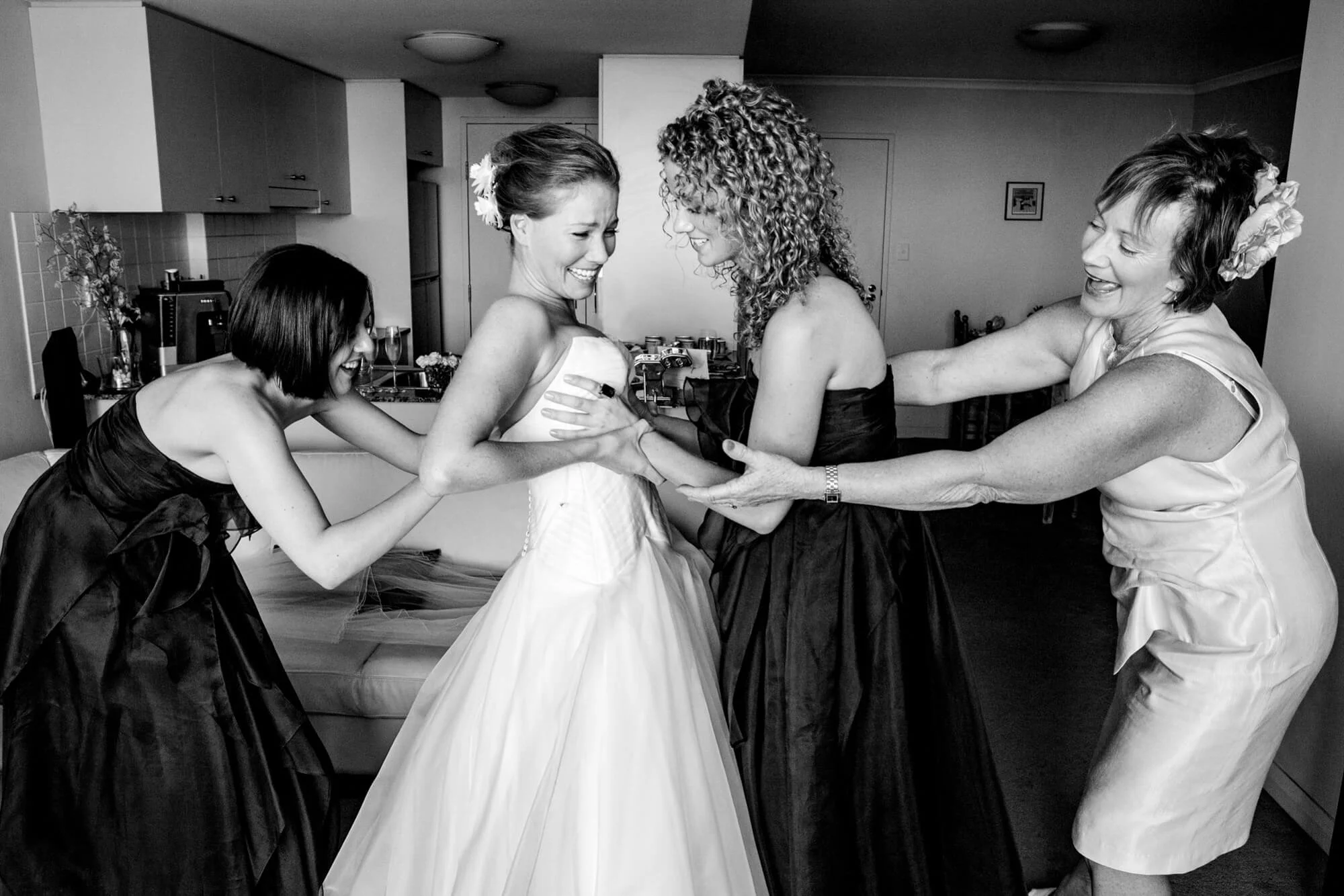 Four women, one in a wedding dress, happily laughing and embracing in a kitchen, dressing room, or home setting.