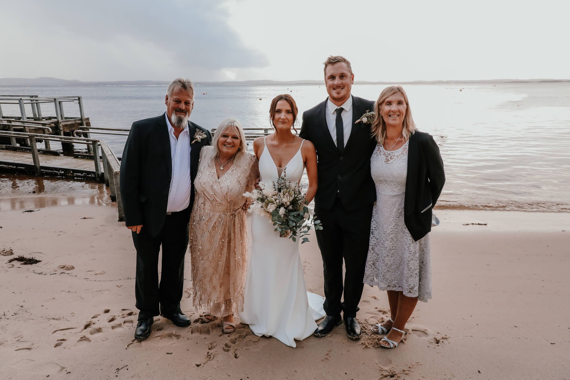 Group of six people in wedding attire posing on a beach near the water, with a cloudy sky overhead.