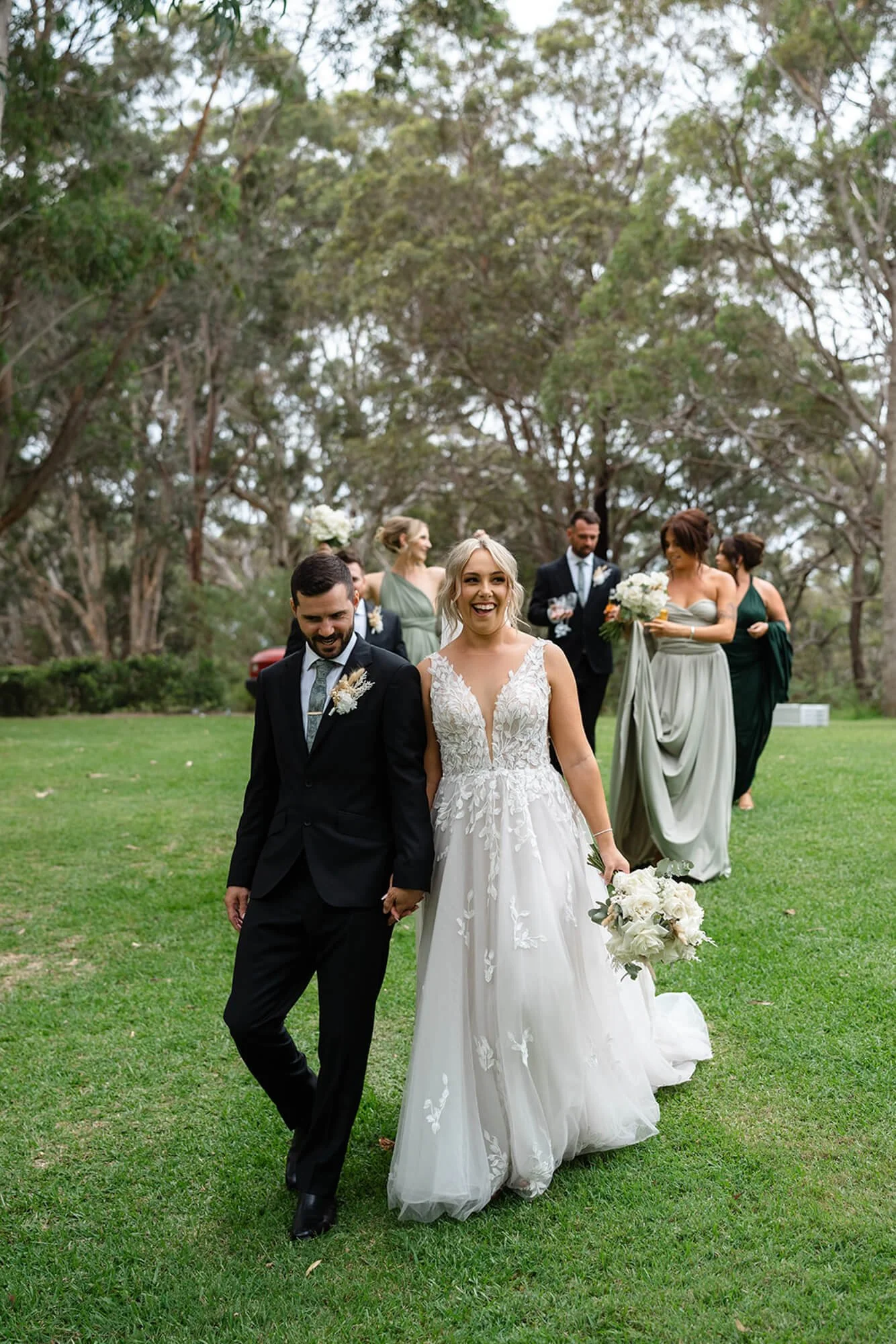 A bride and groom walking hand in hand outdoors, with the bride holding a bouquet of white flowers, surrounded by bridesmaids and groomsmen, in a lush green park with trees.