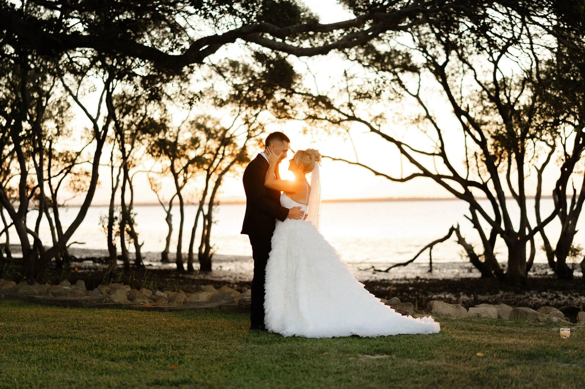 A bride and groom embrace during sunset on a beachside with trees in the background.