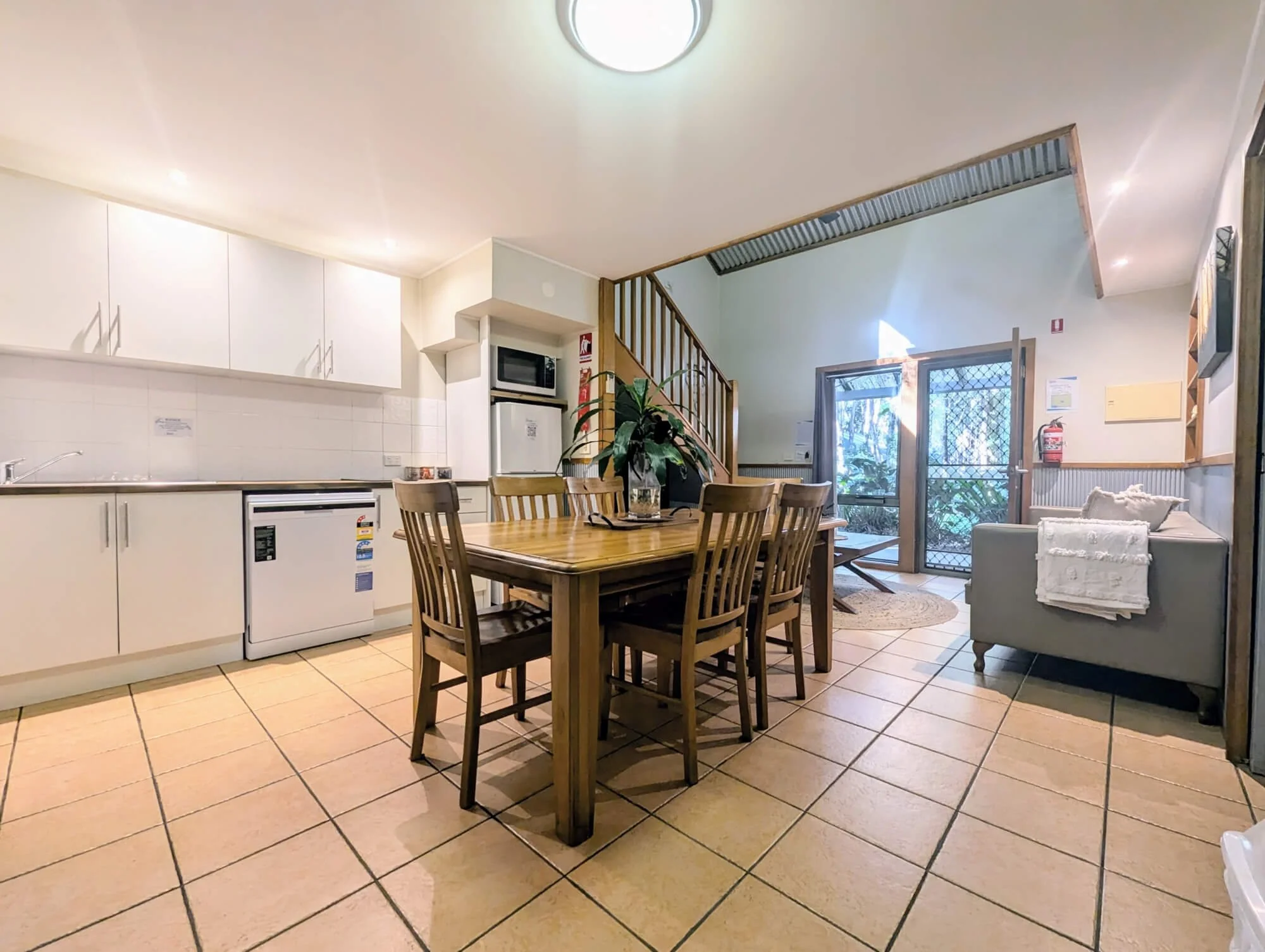 Interior of a dining area with a wooden table and chairs, white kitchen cabinets, a sliding glass door leading to an outside area, and a small gray sofa with a white blanket.