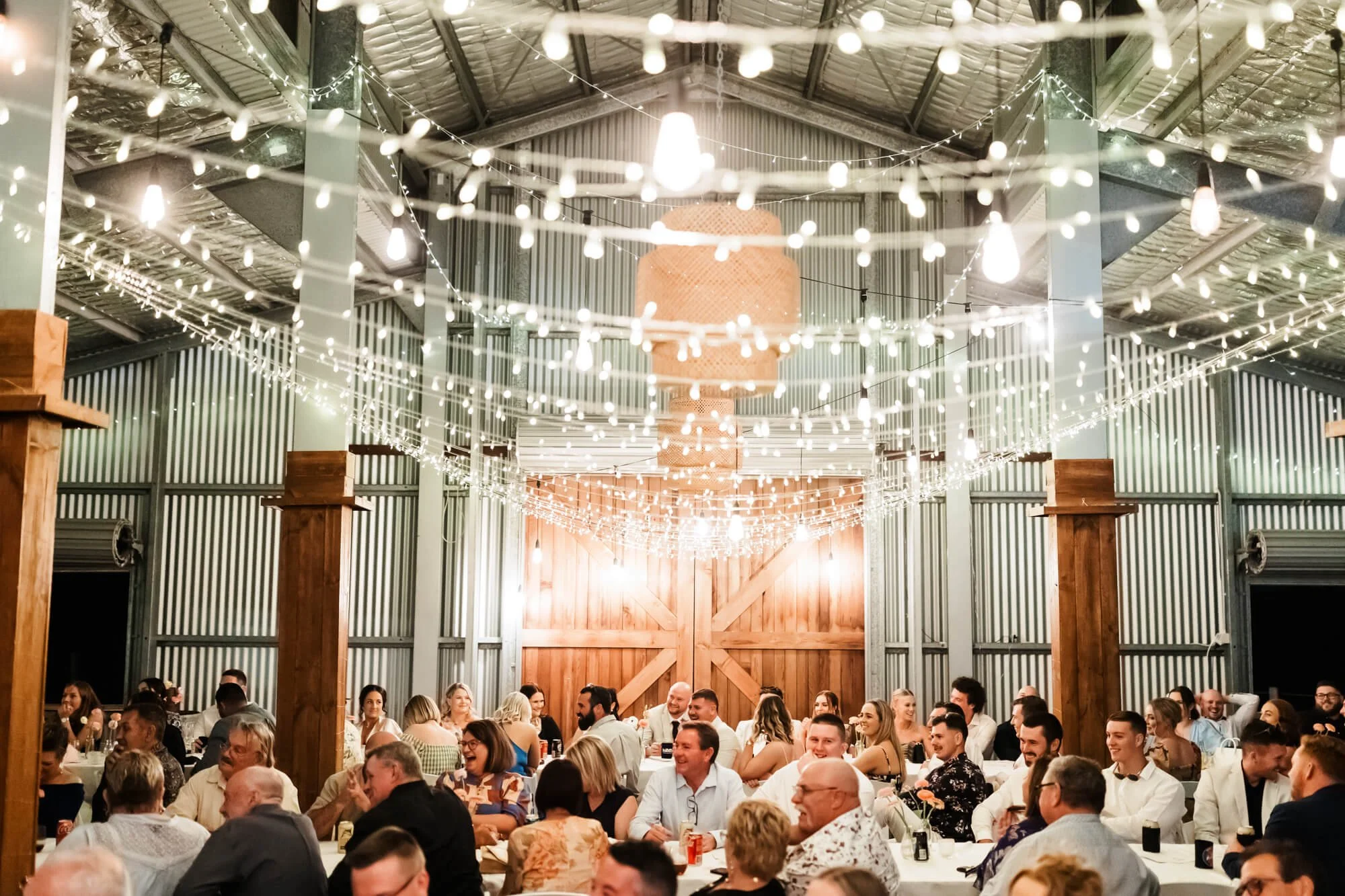 Crowd of people seated at long banquet tables in a decorated barn-style venue with hanging string lights and wooden accents, celebrating an event.