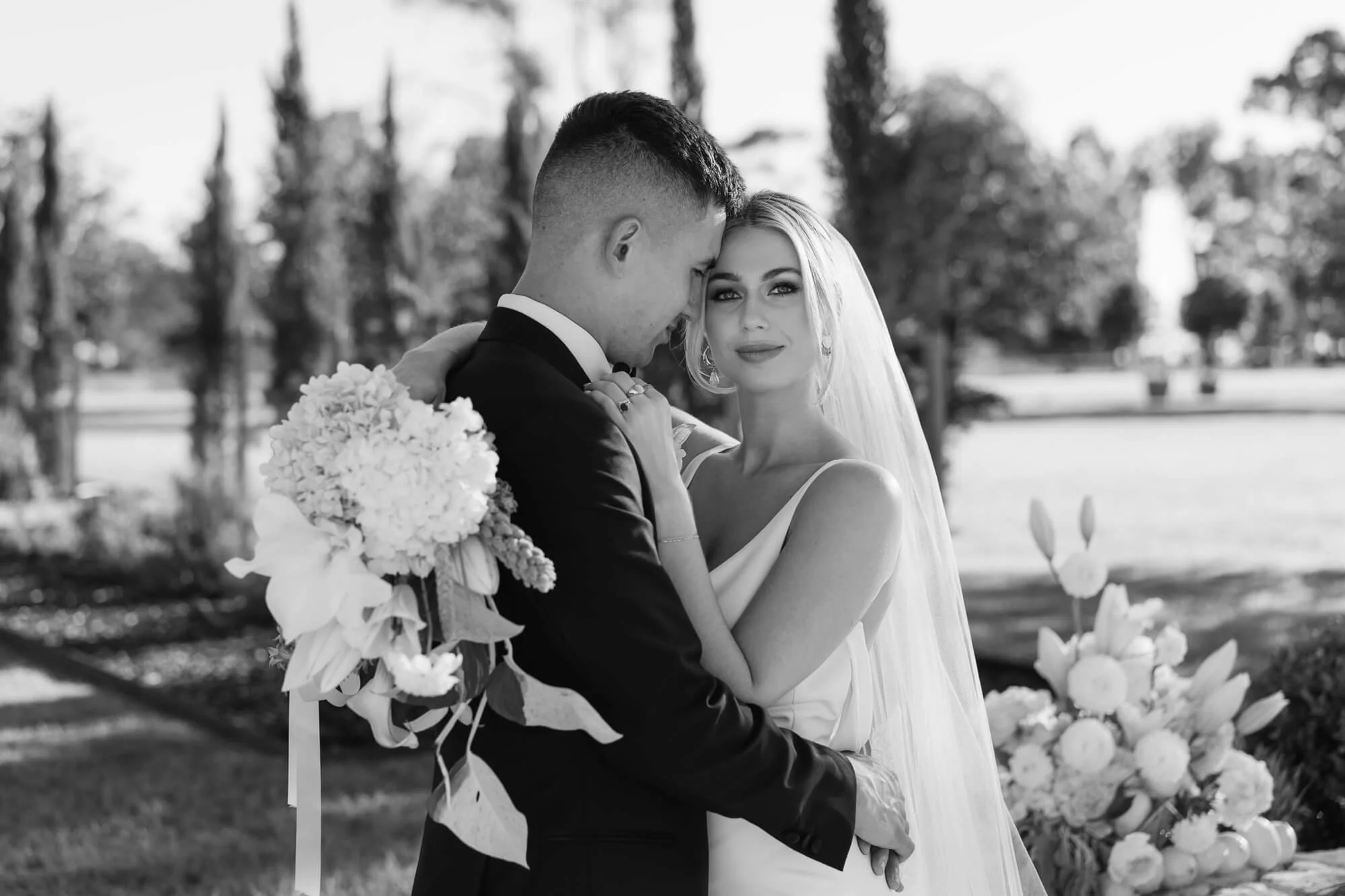 A black and white photograph of a bride and groom embracing outdoors. The bride has blonde hair, wearing a veil and a sleeveless wedding dress, and looks at the camera. The groom, in a suit, holds a bouquet of flowers and leans his forehead against h