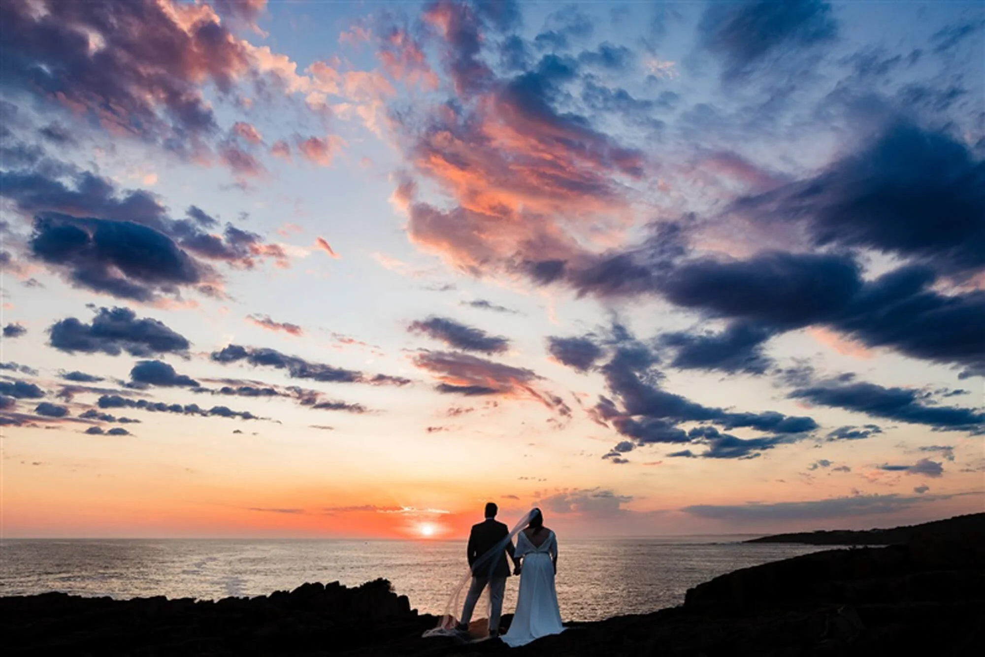 A couple, possibly newlyweds, standing on rocky terrain, holding hands and watching the sunset over the ocean under a colorful sky with clouds.