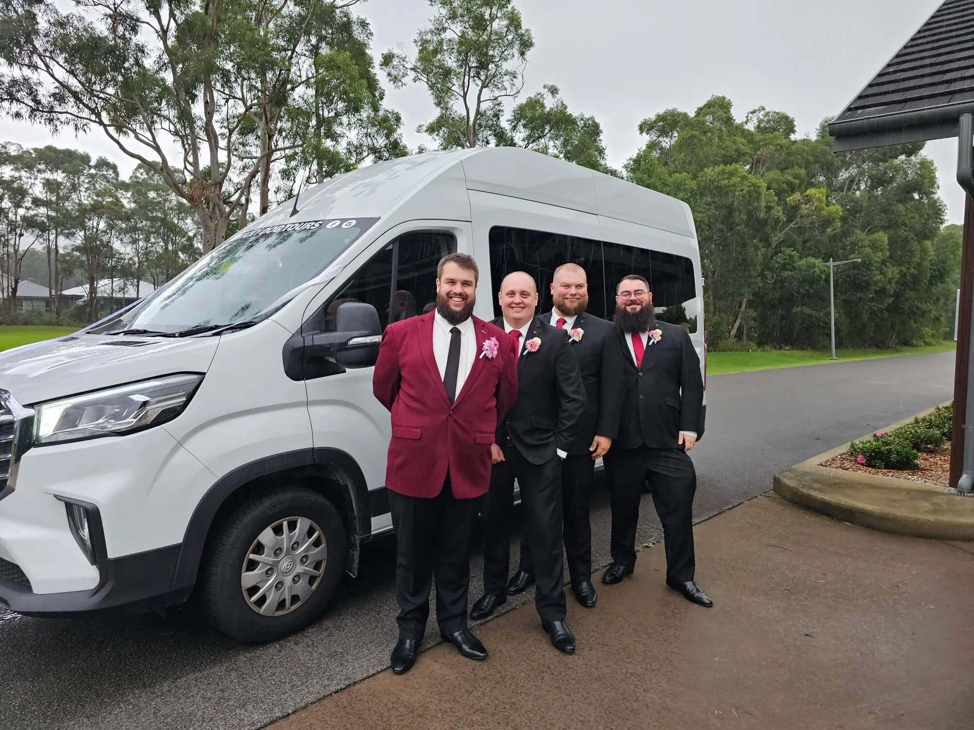 Four men in suits standing in front of a white van. Two of the men are wearing maroon blazers with white boutonnières, and the others are wearing black suits with similar boutonnières. The men are smiling and posing for the photo outdoors, with trees