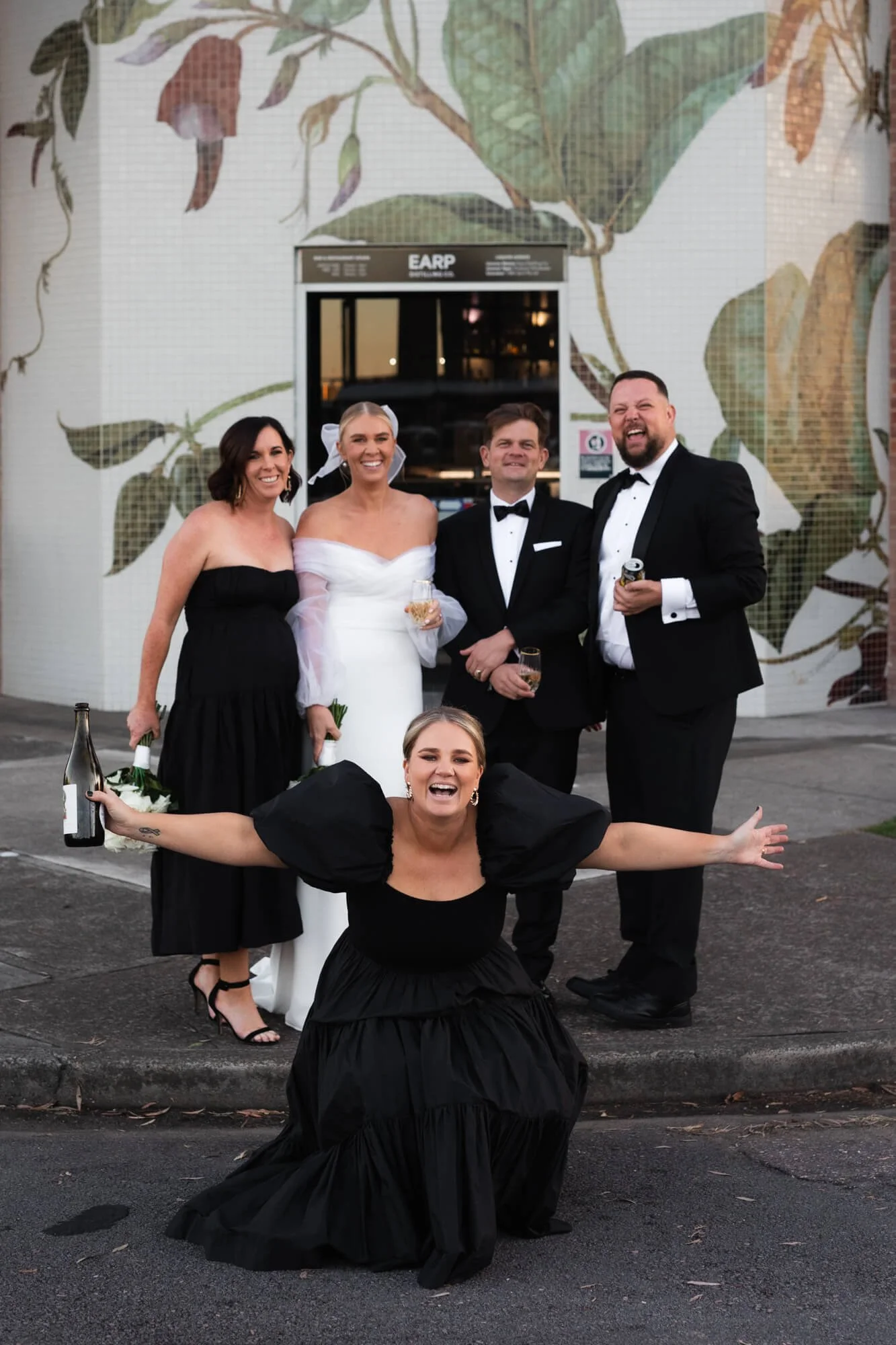 Group of people celebrating at a wedding, five adults, four standing behind, one crouching in front, dressed in formal attire, smiling, holding drinks, sidewalk and mosaic mural with leaf design in the background.