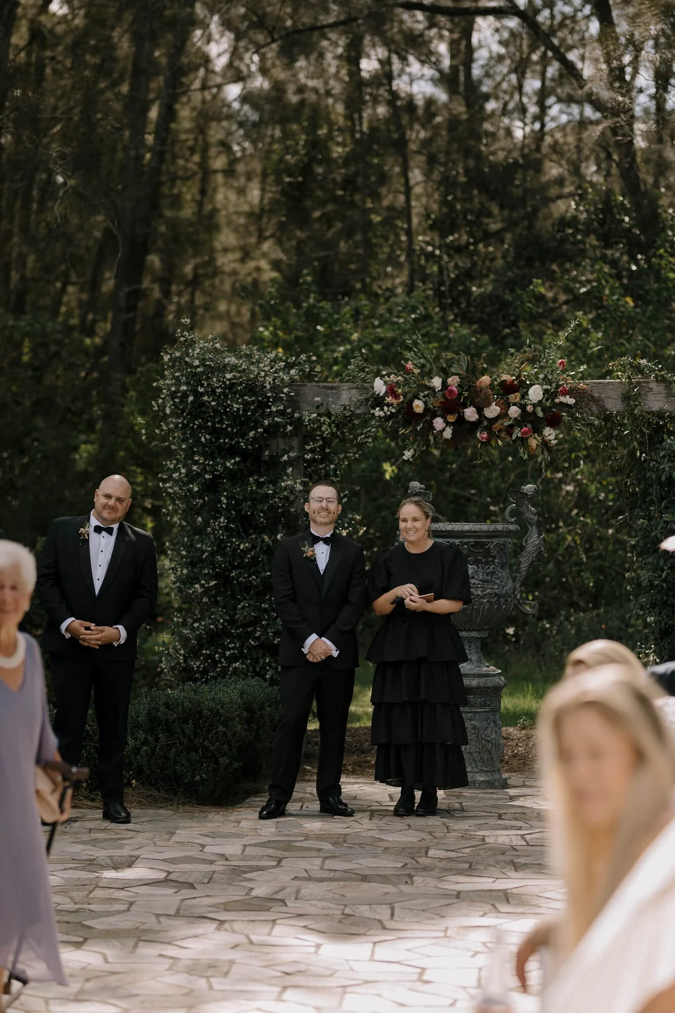 A wedding officiant and two grooms standing outside under a floral arch with trees in the background, during a wedding ceremony.