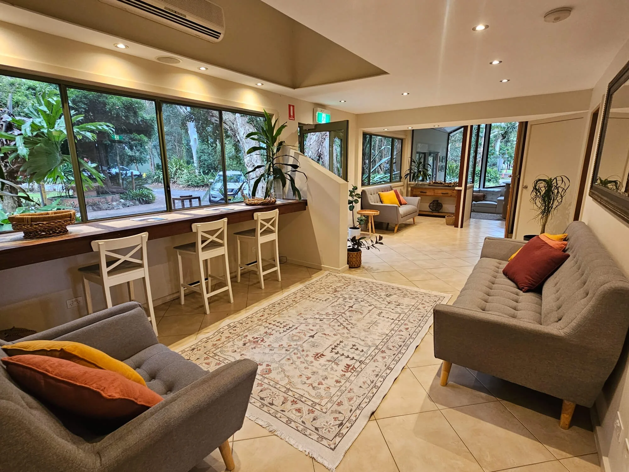 Living room with large windows overlooking greenery, two gray sofas with colorful pillows, a patterned area rug, indoor plants, and a built-in desk with three white chairs.
