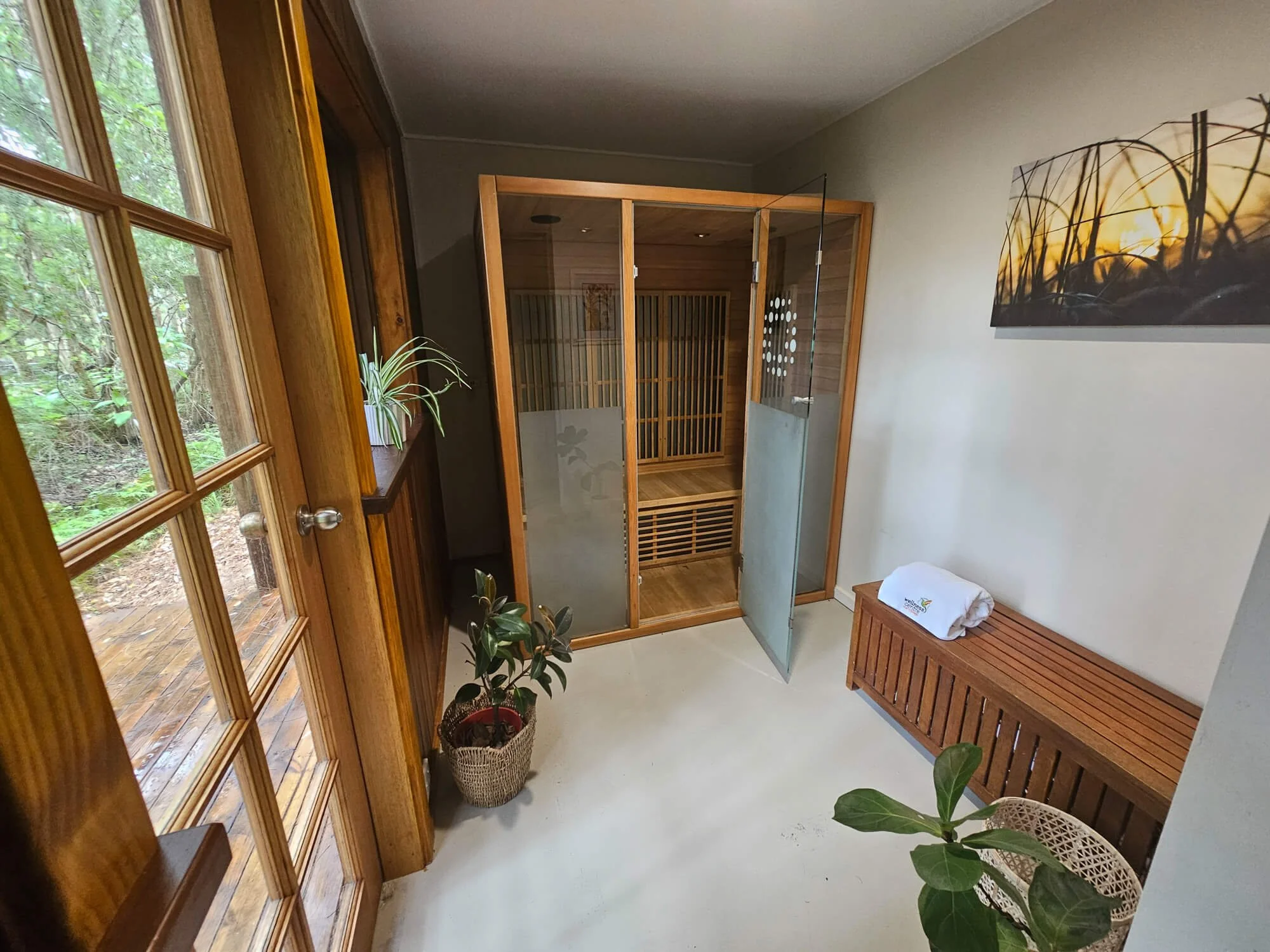 Spa room with wooden sauna, potted plants, a bench with a towel, and a large window showing greenery outside.
