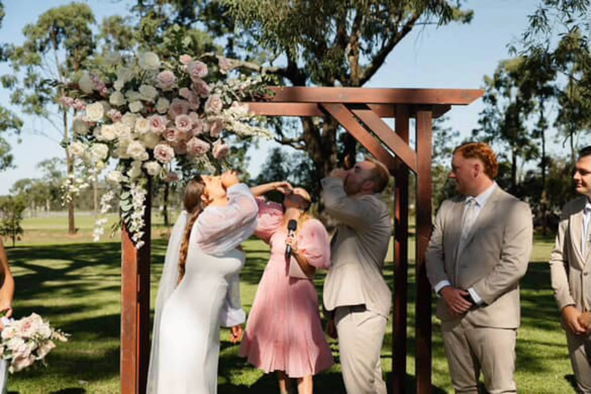 A wedding ceremony outdoors with a bride and groom under a wooden arch decorated with pink and white flowers, with people celebrating around them.