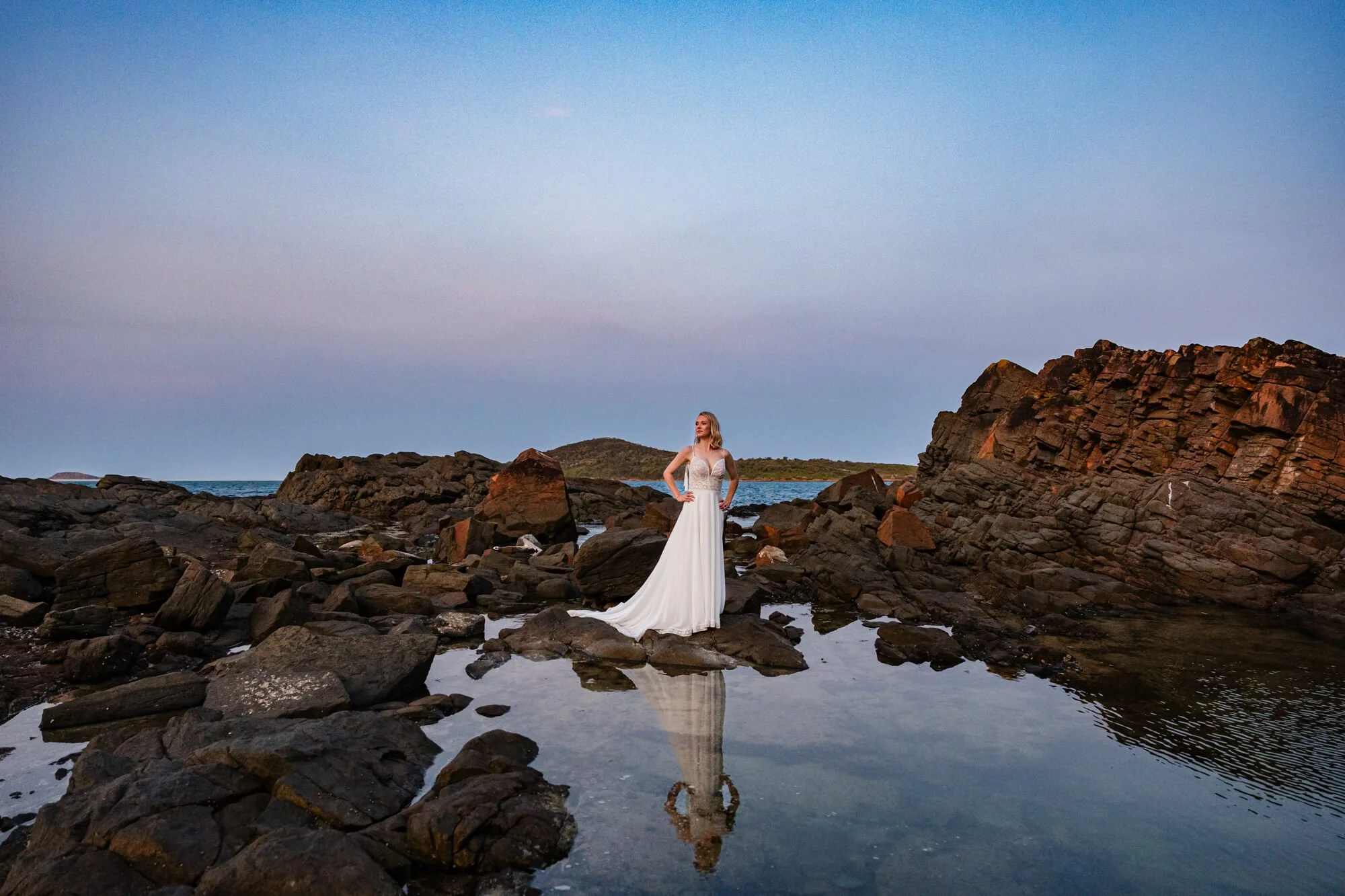 A woman in a white wedding dress standing among rocks near the shore, with a reflection in a small pool of water, during a sunset or dusk with a clear sky and distant land in the background.