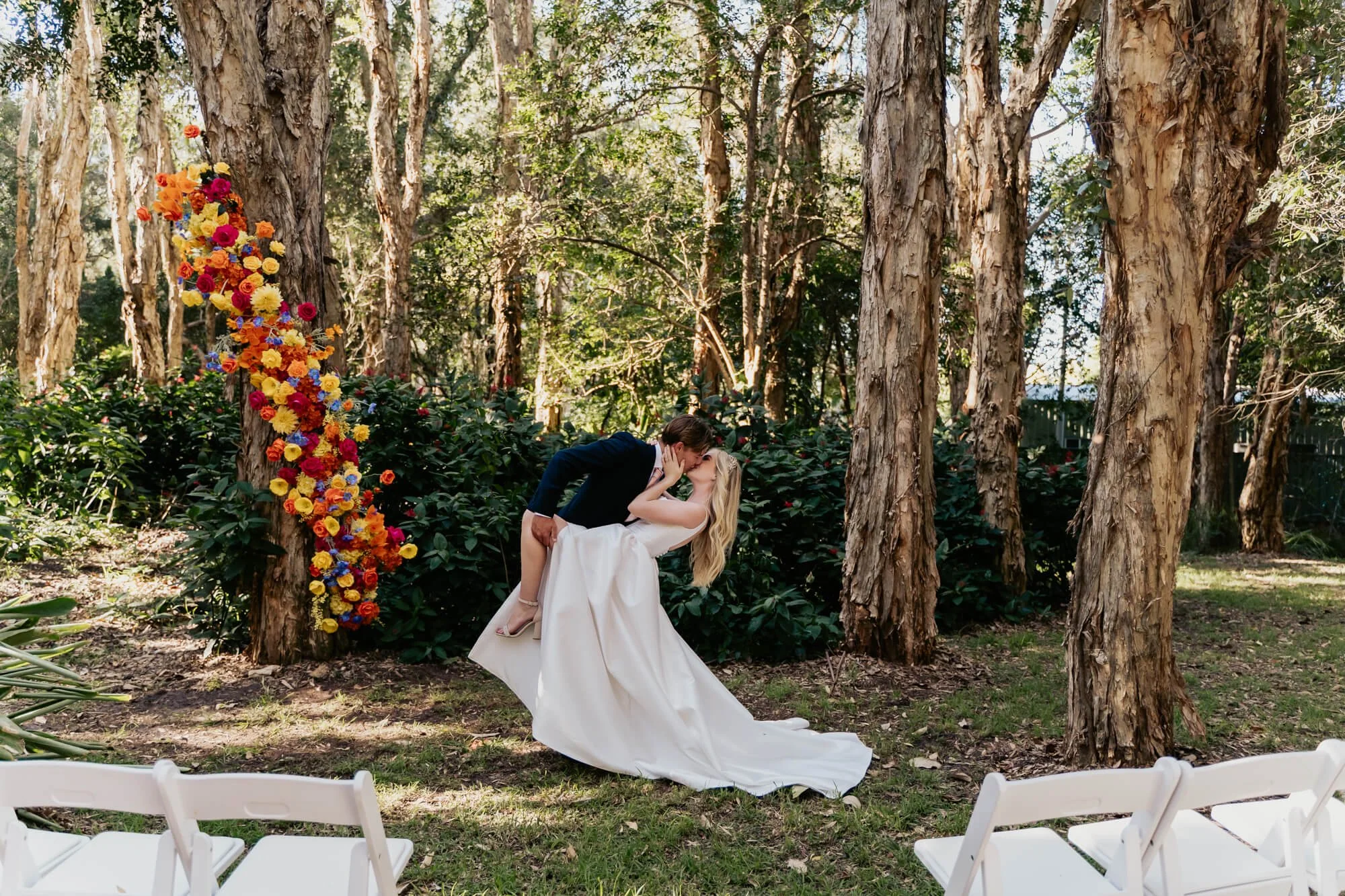 A couple in wedding attire sharing a kiss in a forest setting with trees and colorful flowers, and white chairs in the foreground.