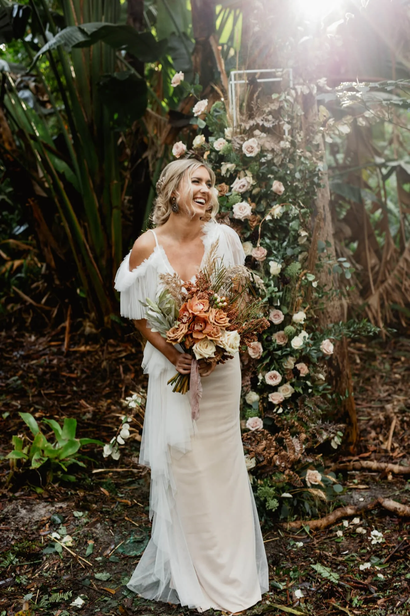 A smiling bride in a white wedding dress holding a bouquet of peach, white, and green flowers. She is standing outdoors among lush greenery and flowers, with sunlight shining through the trees.