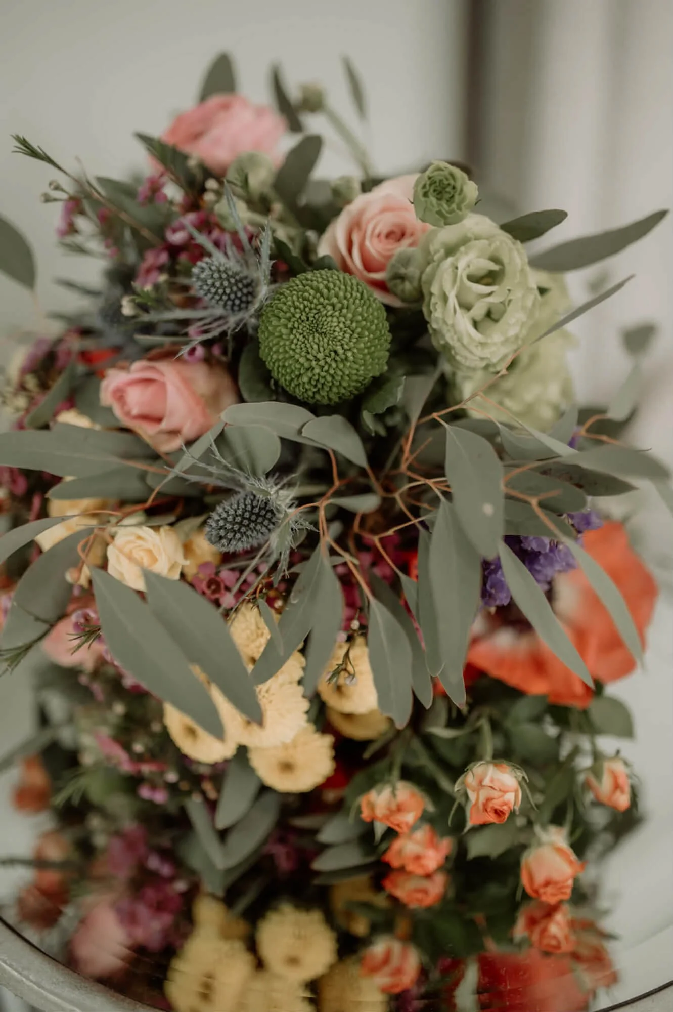 A close-up photo of a mixed flower bouquet with pink roses, green pom-poms, purple and peach flowers, and eucalyptus leaves.