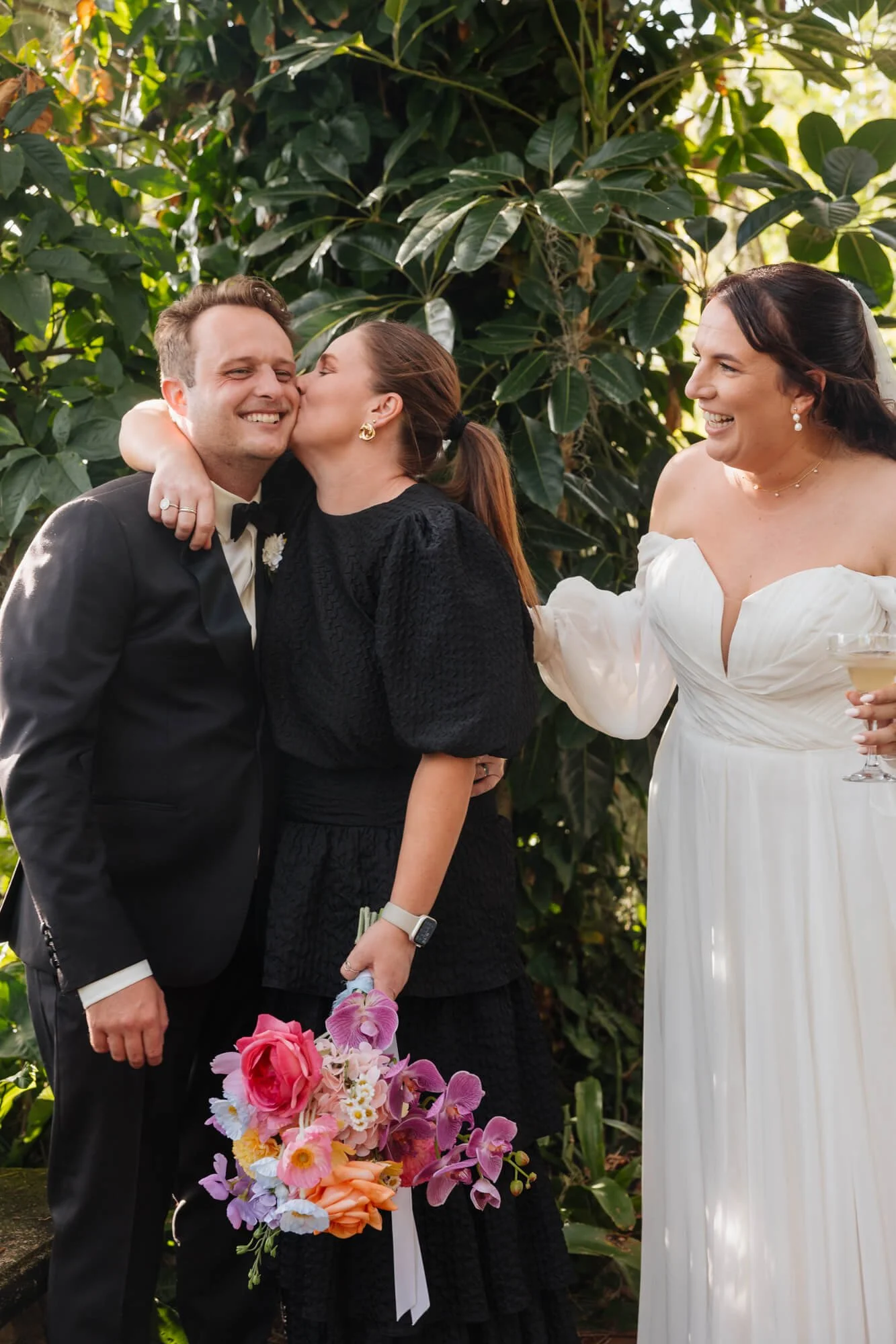 A wedding celebration showing a woman in a white dress giving a kiss on the cheek to a man in a black suit, with another smiling woman in a black dress holding a bouquet of flowers nearby.
