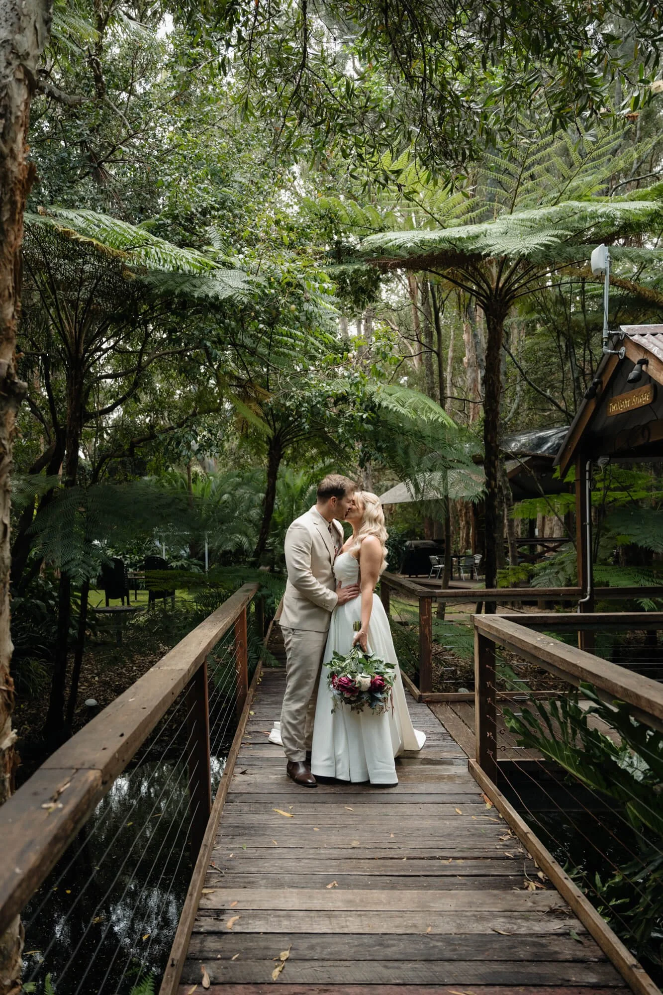 A bride and groom sharing a kiss on a wooden walkway in a lush green forest, surrounded by large leafy plants and trees.