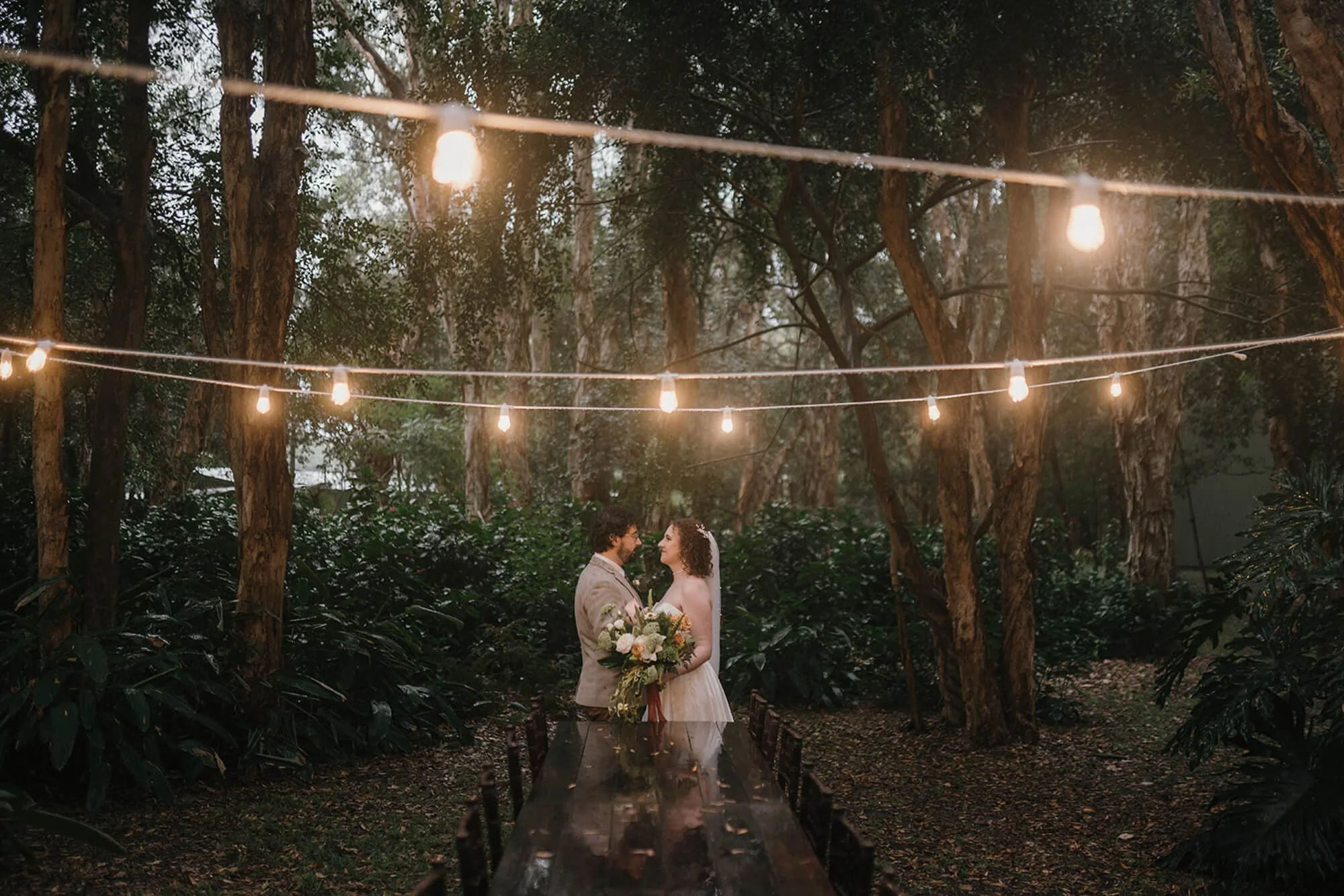 A bride and groom standing close together under string lights in a wooded outdoor setting, sharing a moment.
