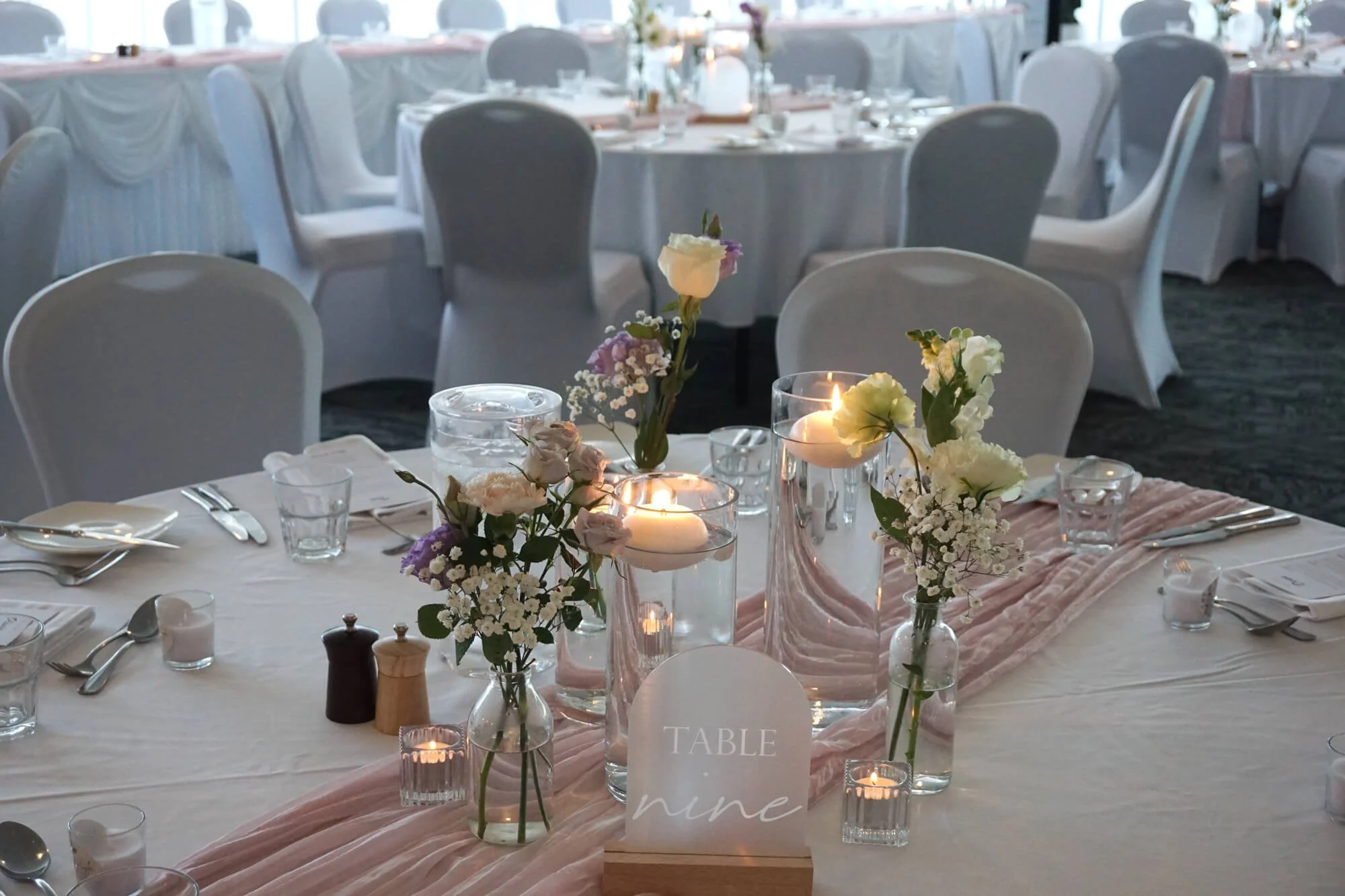 Elegant banquet table decorated with pastel pink and white flowers in glass vases, floating candles, and a pink table runner. White plates, utensils, and glasses are arranged around the table, with a table sign reading 'TABLE nine.' In the background