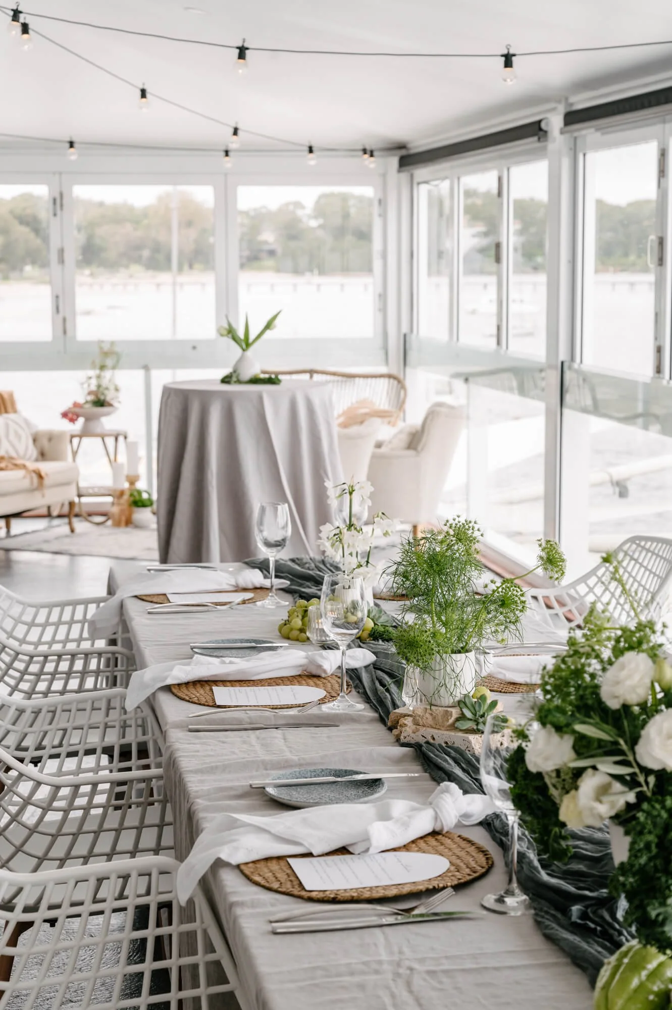 Elegant dining setup inside a bright, modern room with large windows overlooking a water body, decorated with white flowers and greenery, set for a formal meal.